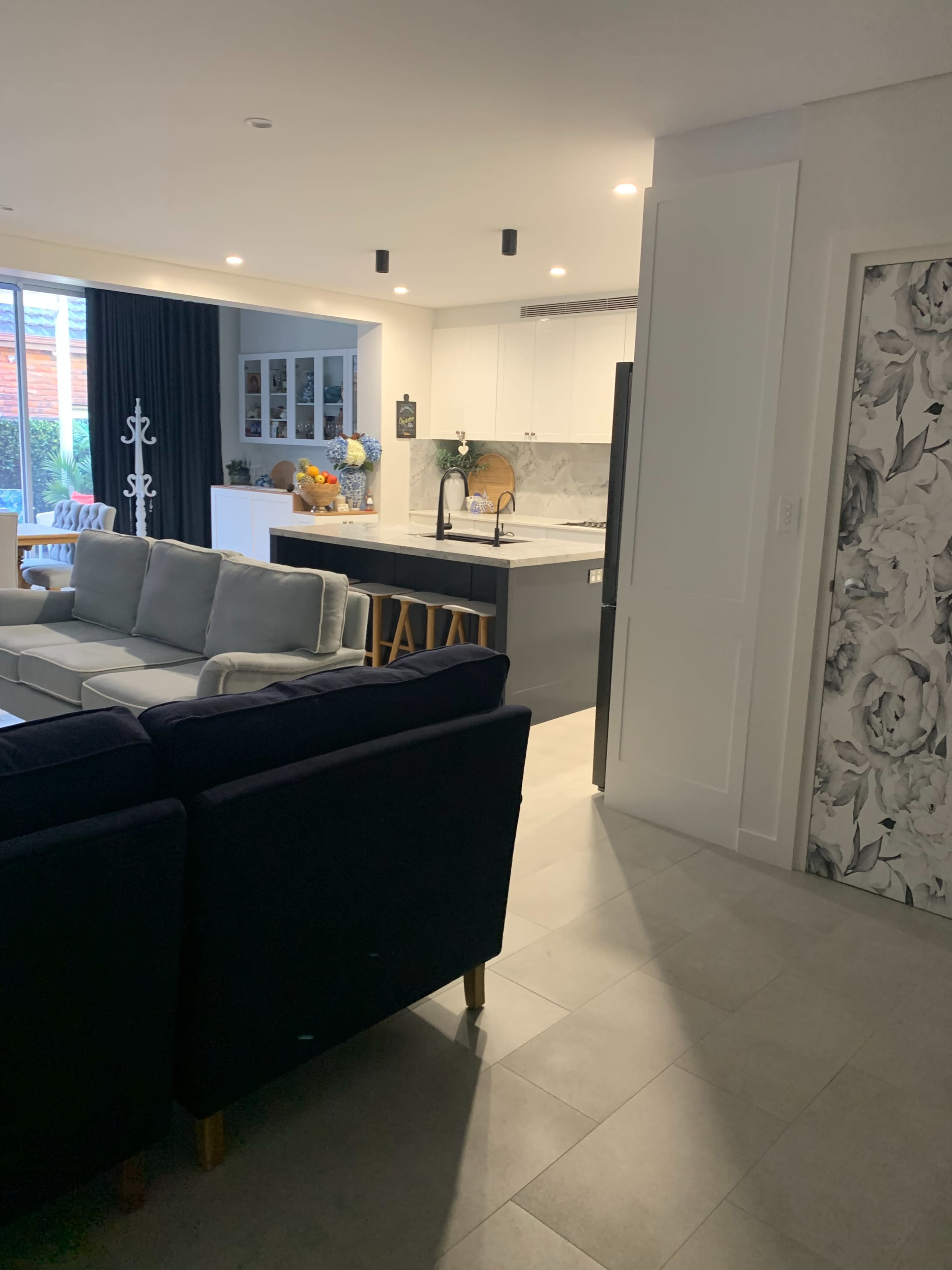 The image shows a modern open-concept living area leading into a kitchen, featuring a seating area with a sectional sofa and a dining space with bar stools.