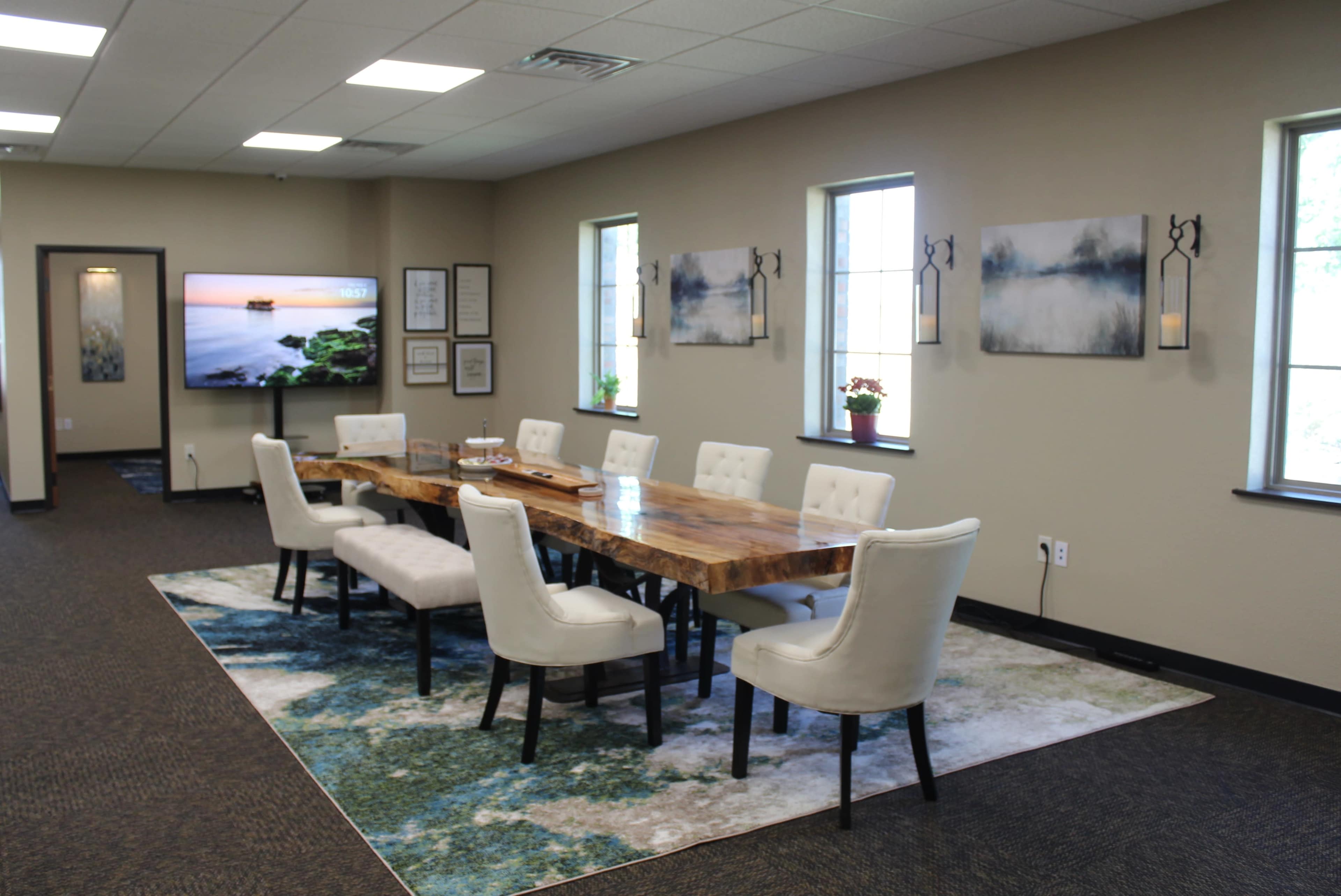 A long wooden table with white upholstered chairs is set in a well-lit meeting room, featuring windows and a large television screen on one wall.