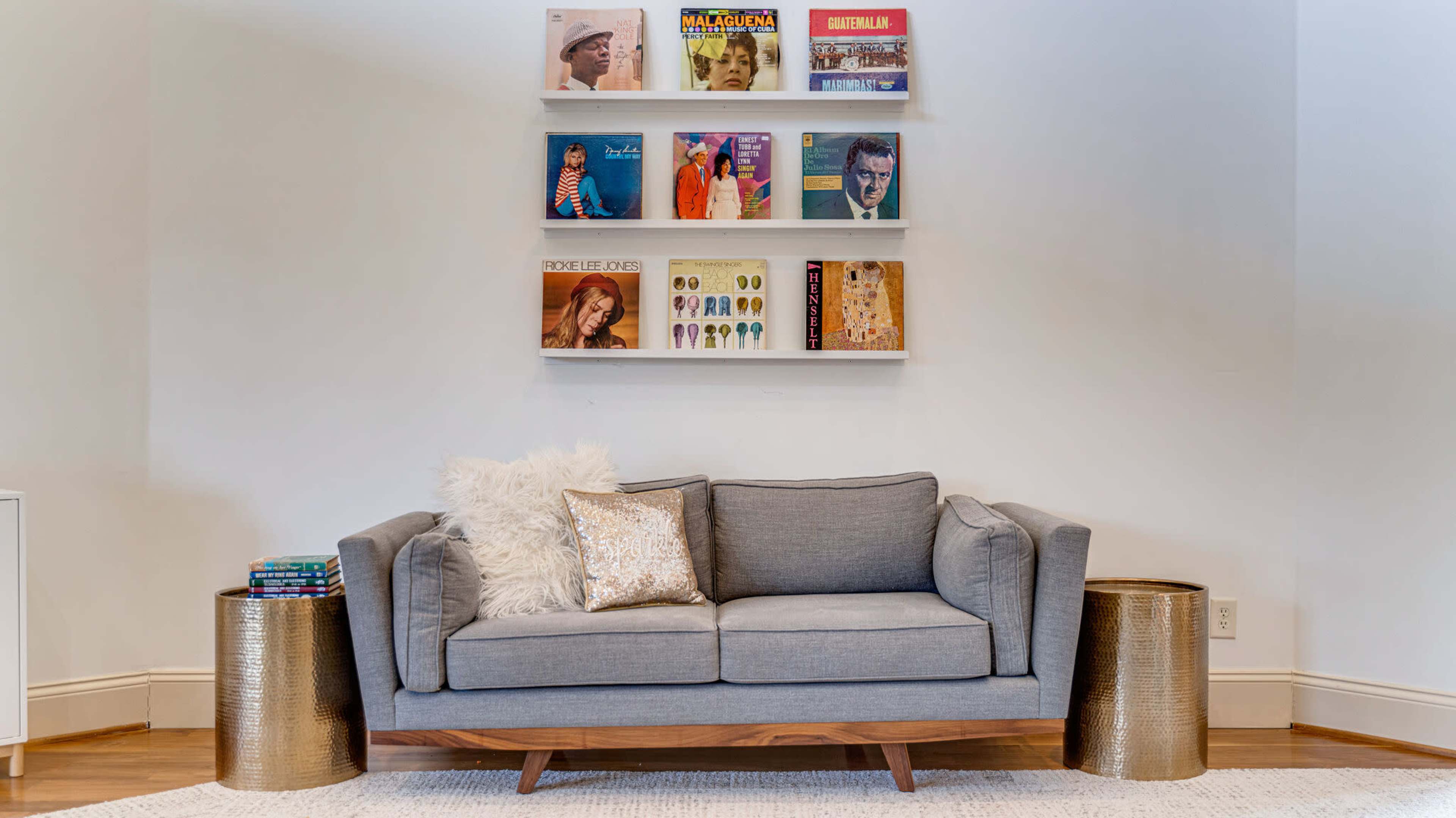 A gray sofa with decorative pillows sits in front of a wall-mounted shelf displaying various vinyl records, alongside two metallic side tables.