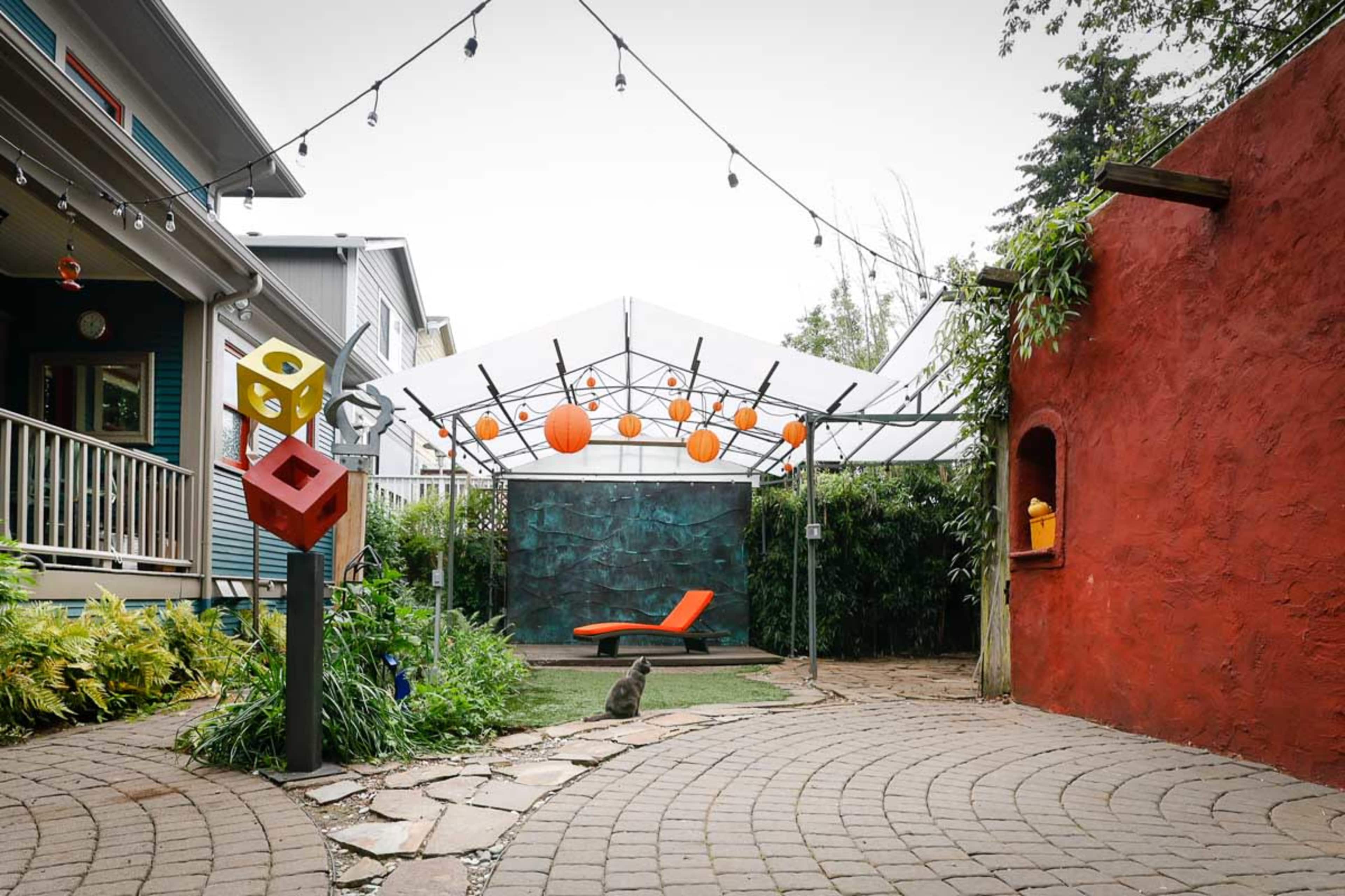 A landscaped outdoor space featuring a patio with a geometric sculpture, a red wall, an orange lounge chair, and string lights overhead.