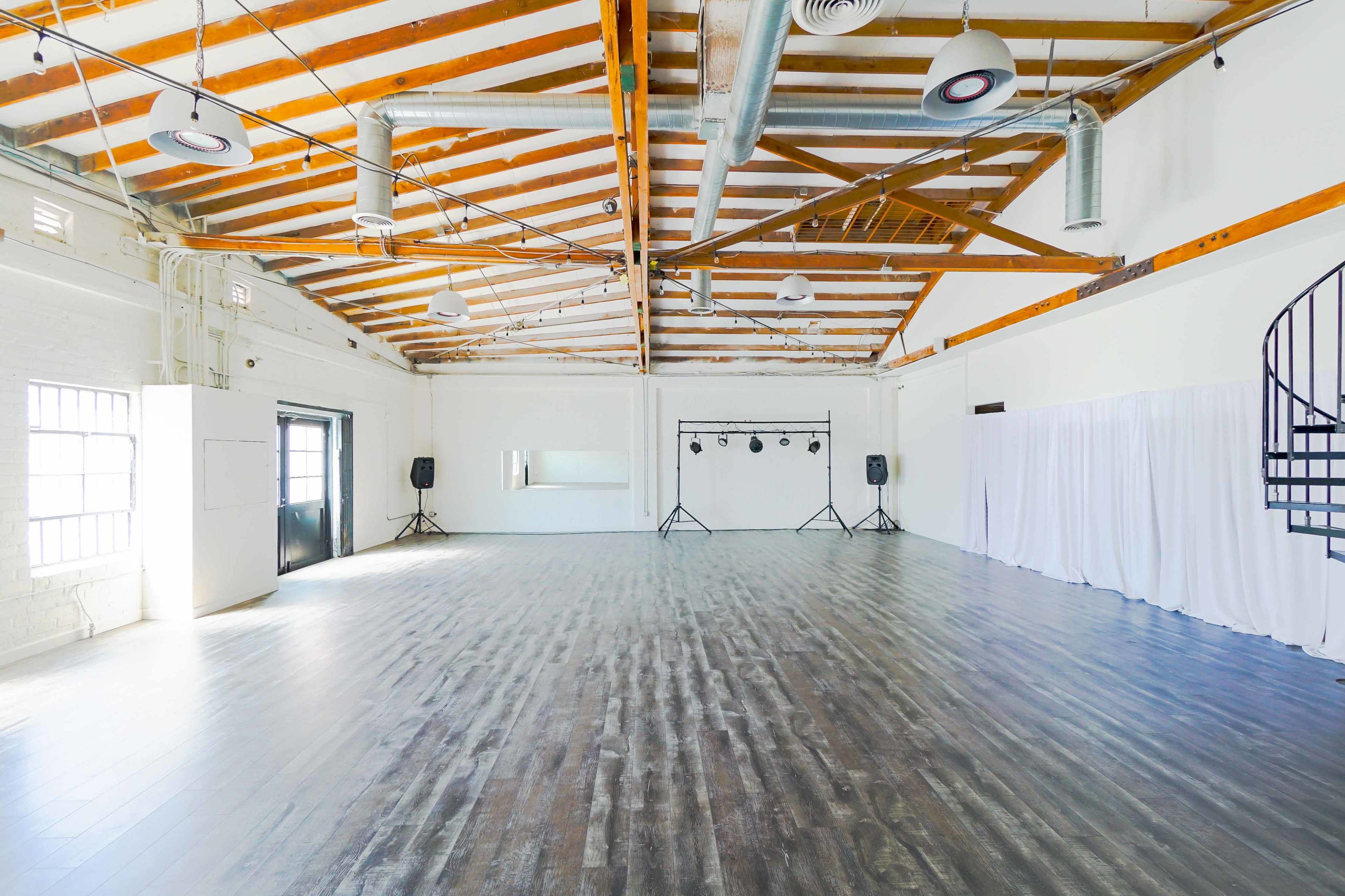 A spacious, brightly lit industrial-style room with wooden beams, a polished wooden floor, and minimal furnishings, including a mirror and audio equipment.