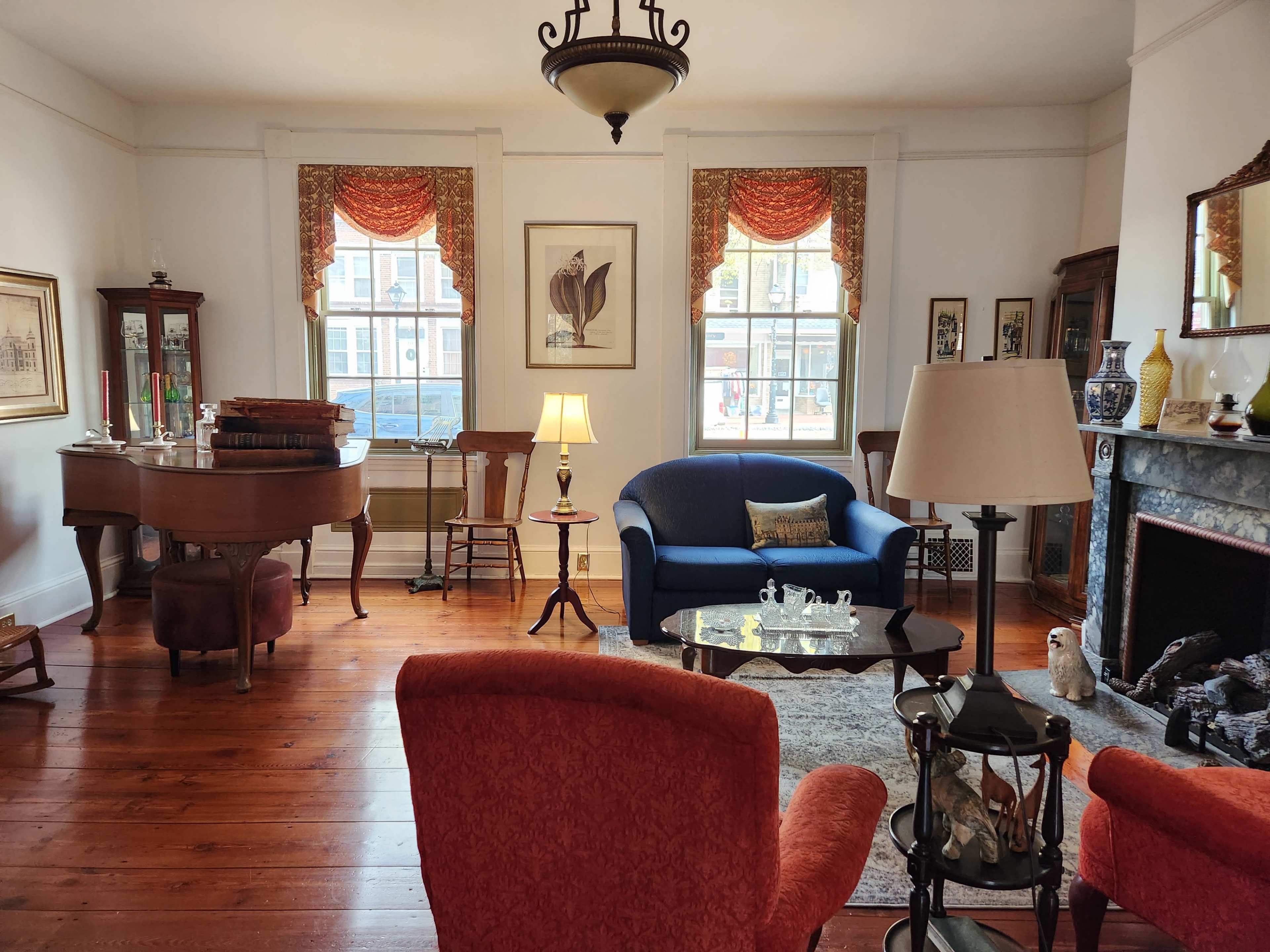 A well-furnished living room with a piano, a blue sofa, and a coffee table, set against wooden flooring and bright windows.