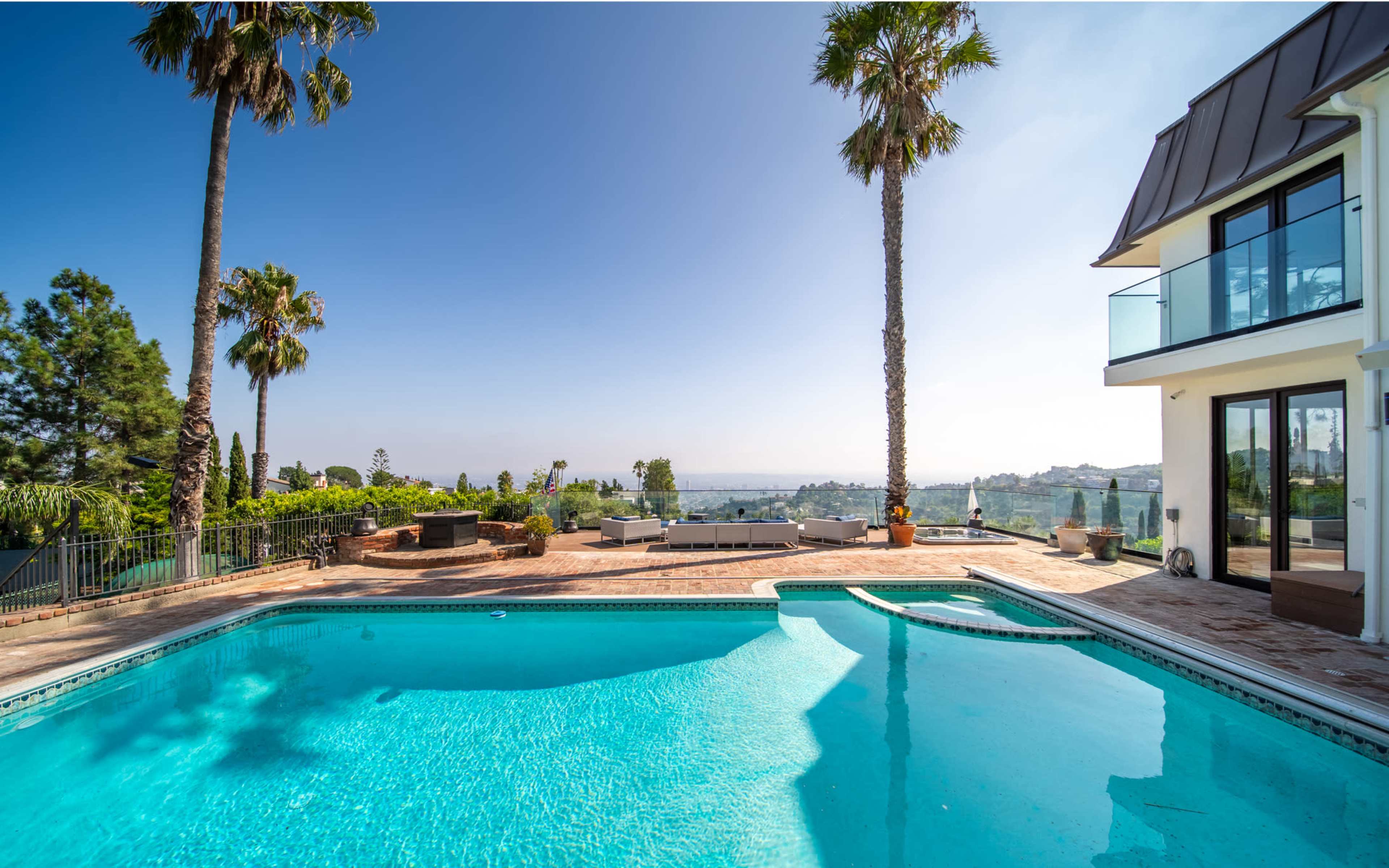 The image shows a clear swimming pool surrounded by palm trees and a view of distant hills under a blue sky.