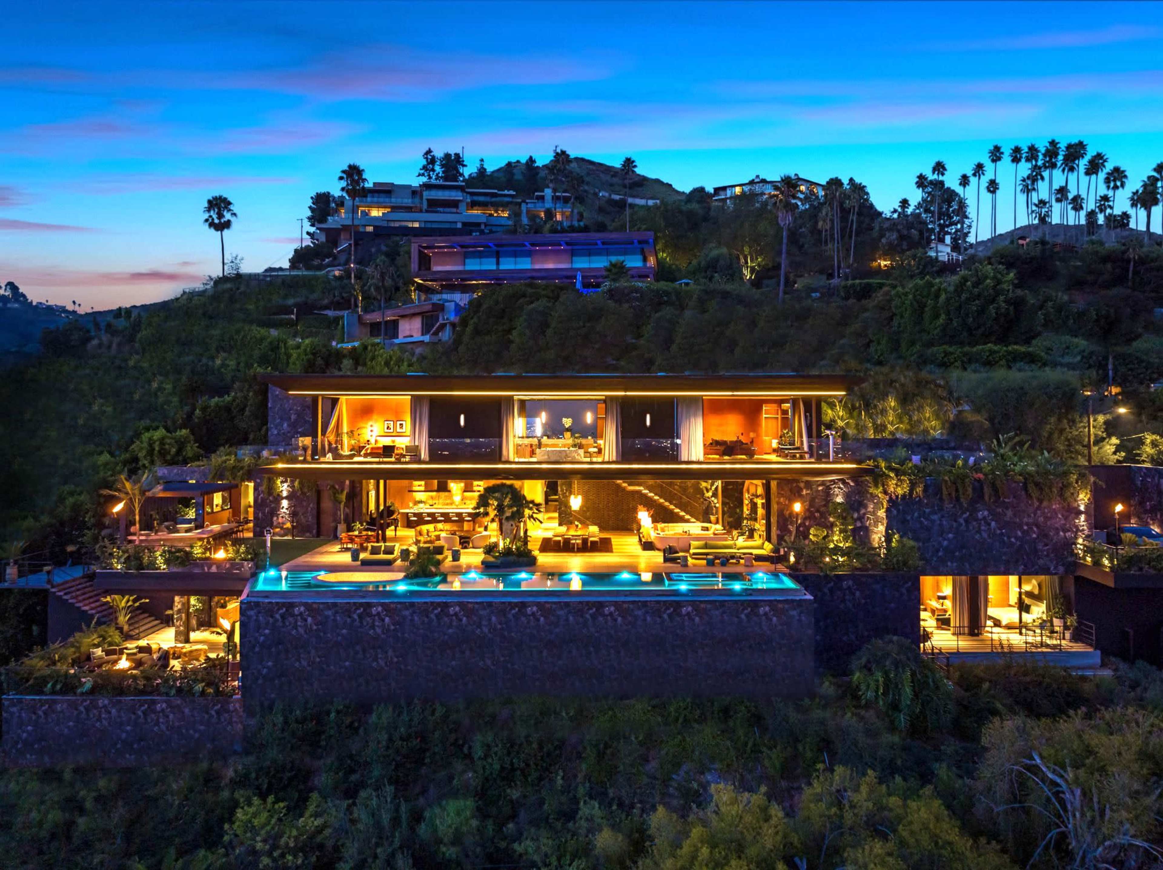 The image shows a modern hillside villa featuring multiple levels, large windows, and a pool illuminated by lights under a twilight sky.