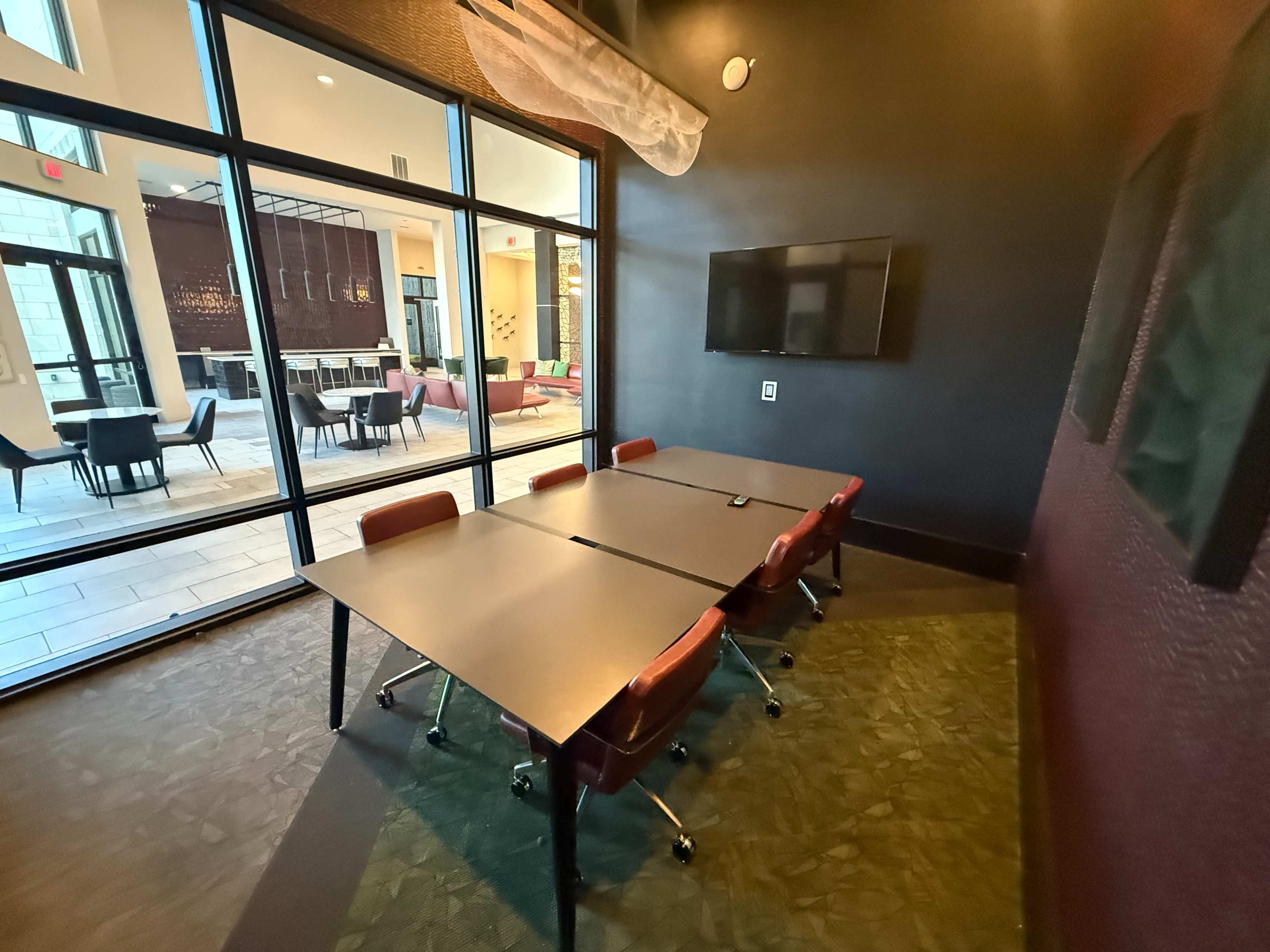 The image shows a modern conference room with a large table and red chairs, featuring large windows that overlook a communal area.