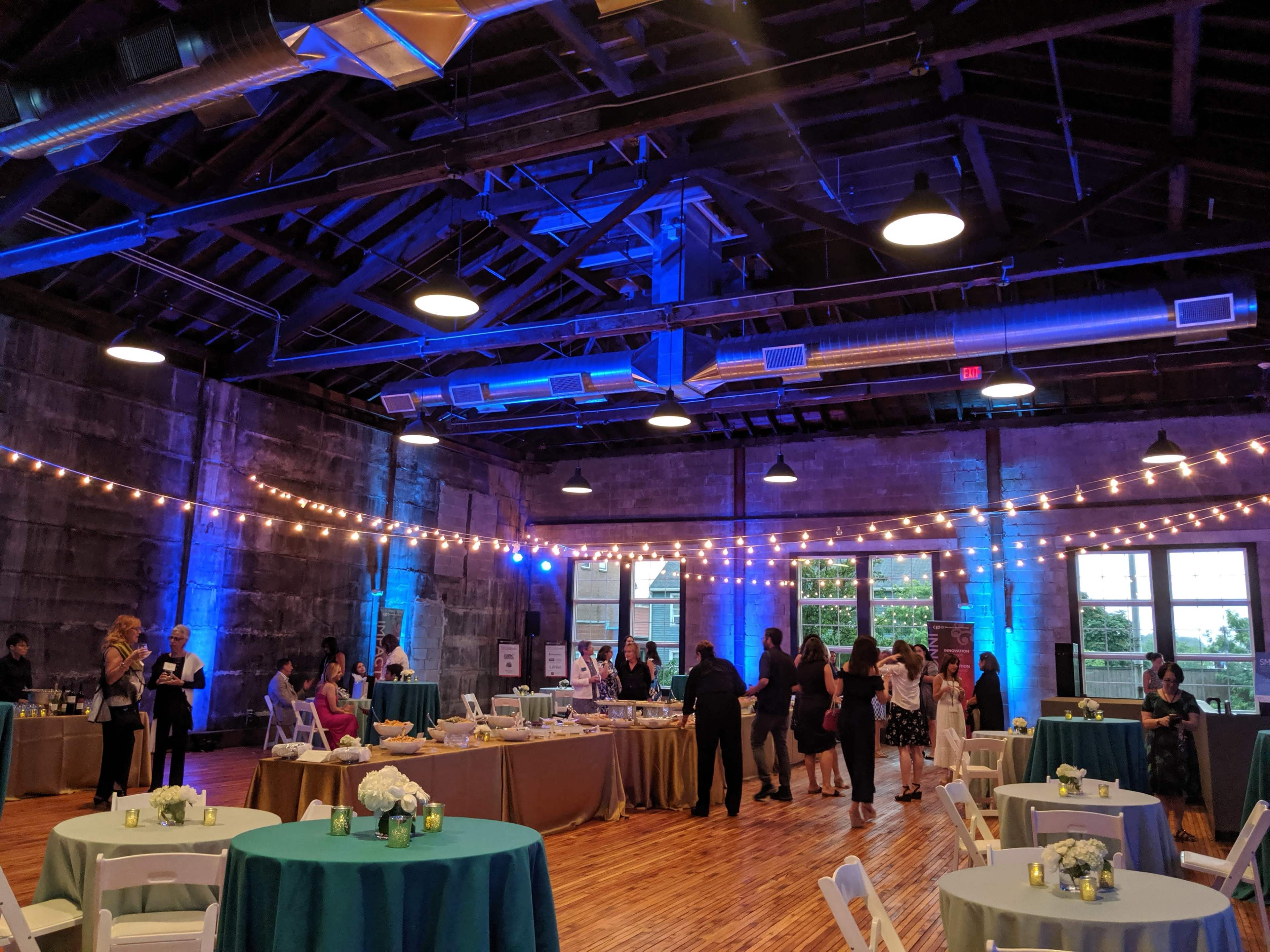 A spacious event venue features round tables with blue tablecloths, food stations, and string lights overhead, as guests mingle and socialize.