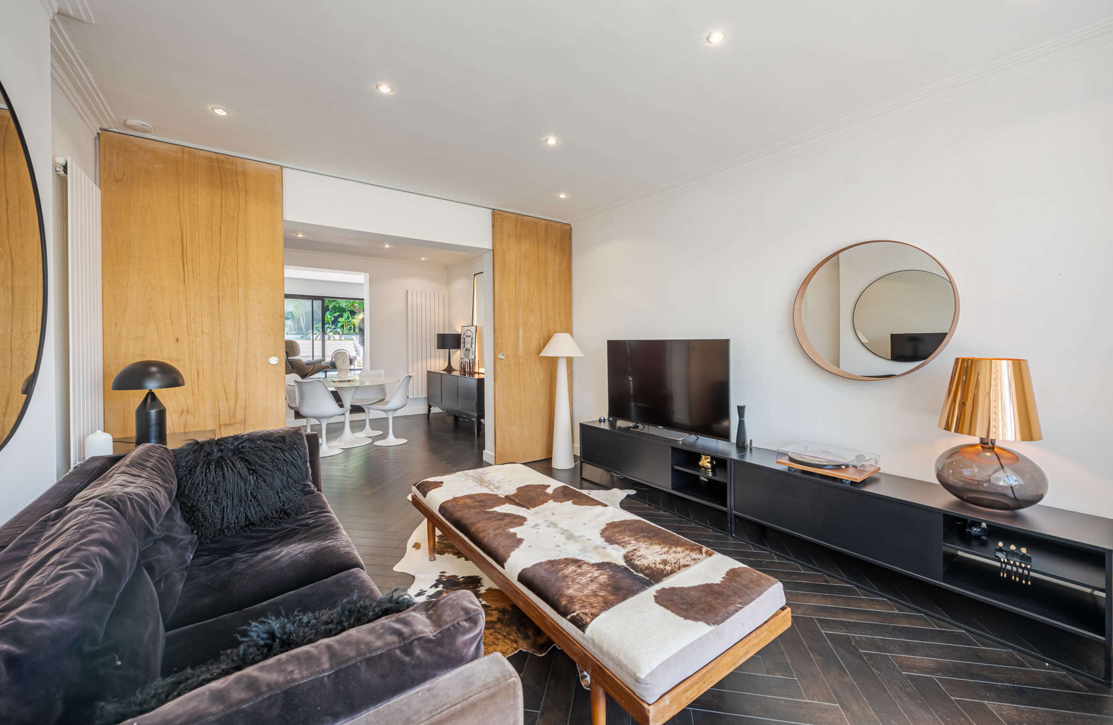 The image shows a modern living room with a dark sofa, a coffee table, a television on a black media unit, and a large round mirror on the wall.