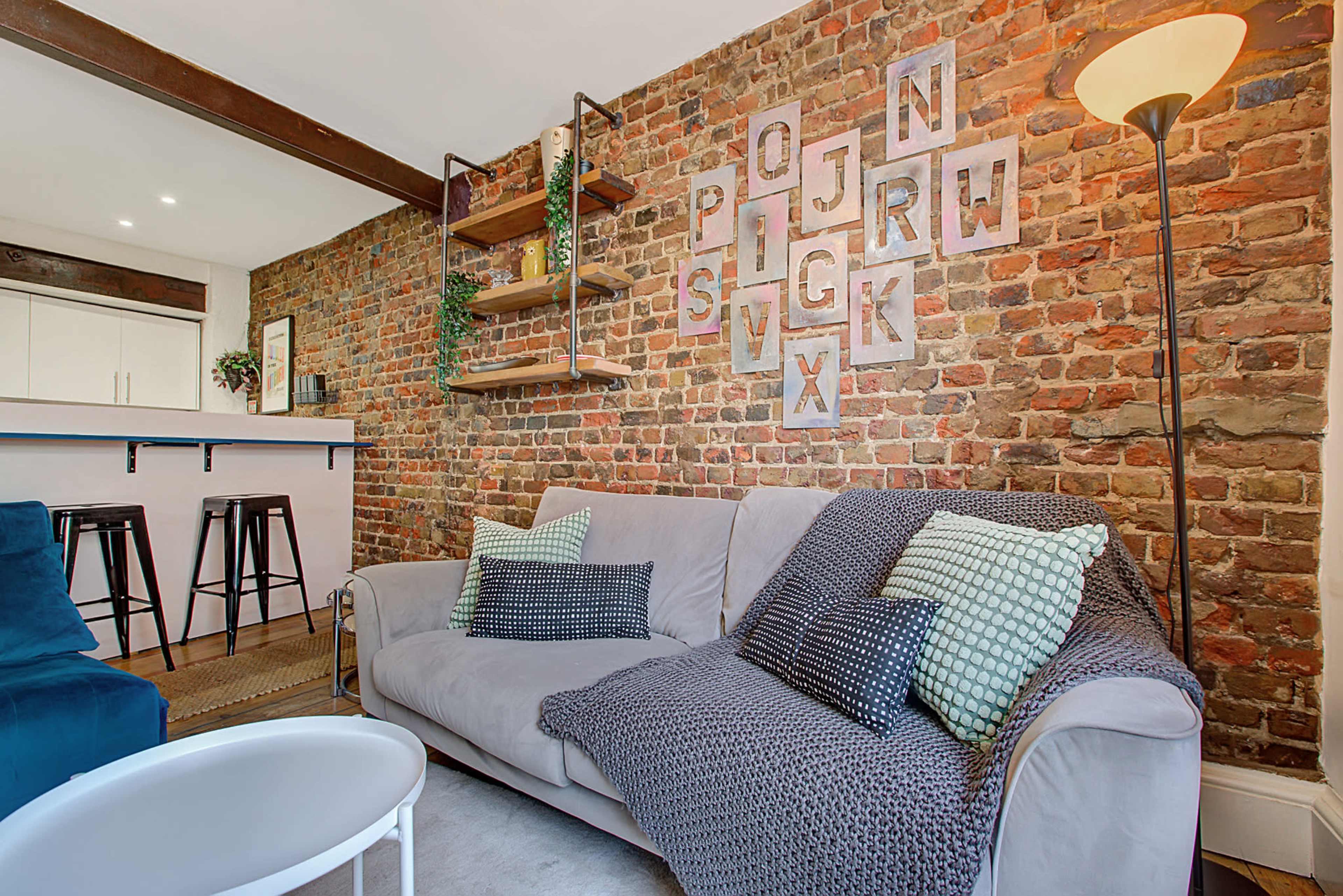 A cozy living room features a gray sofa with patterned cushions, a brick wall, and wooden shelving displaying decorative items.