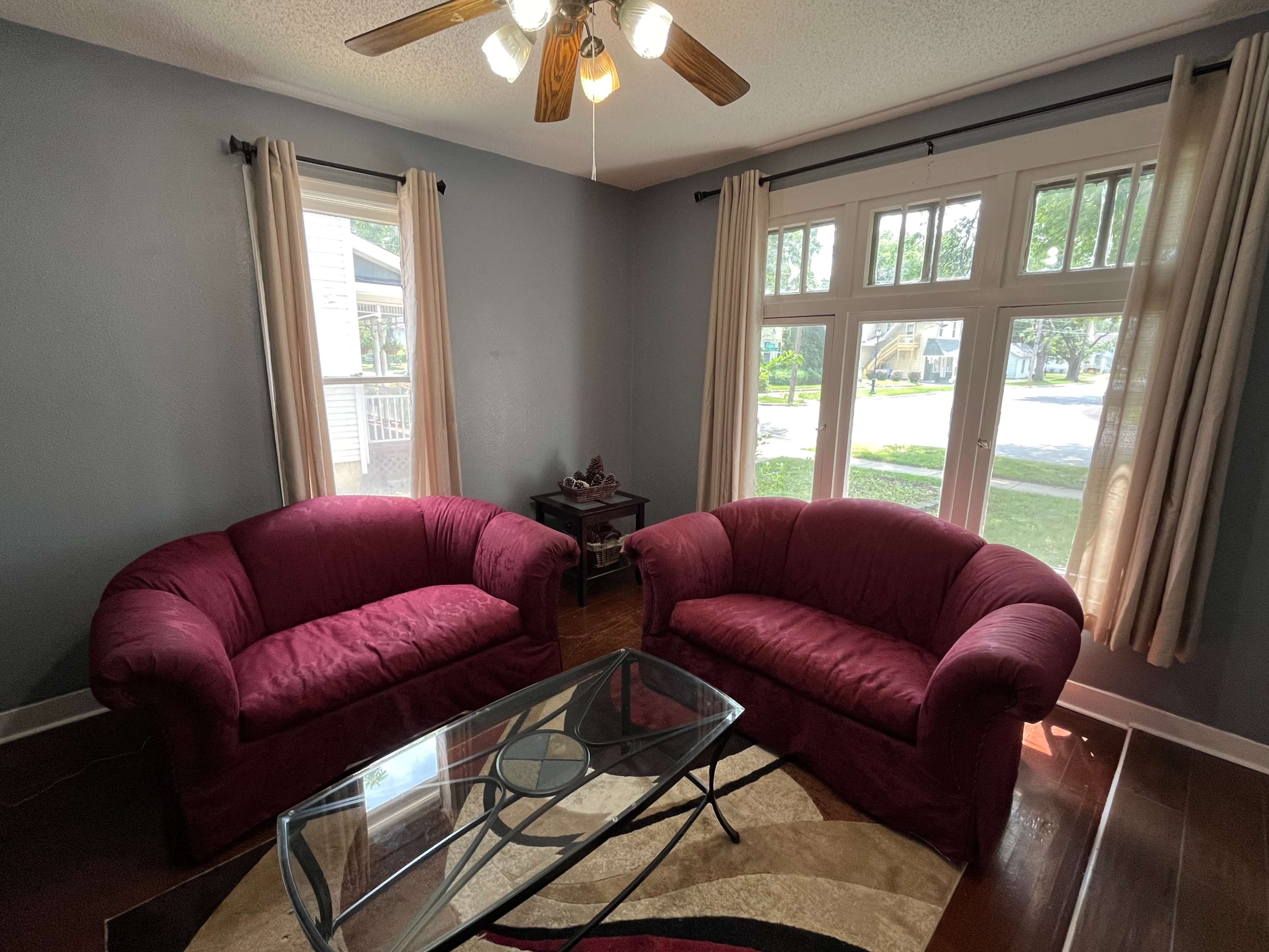 A living room with two red upholstered chairs, a glass coffee table, and large windows allowing natural light to enter.