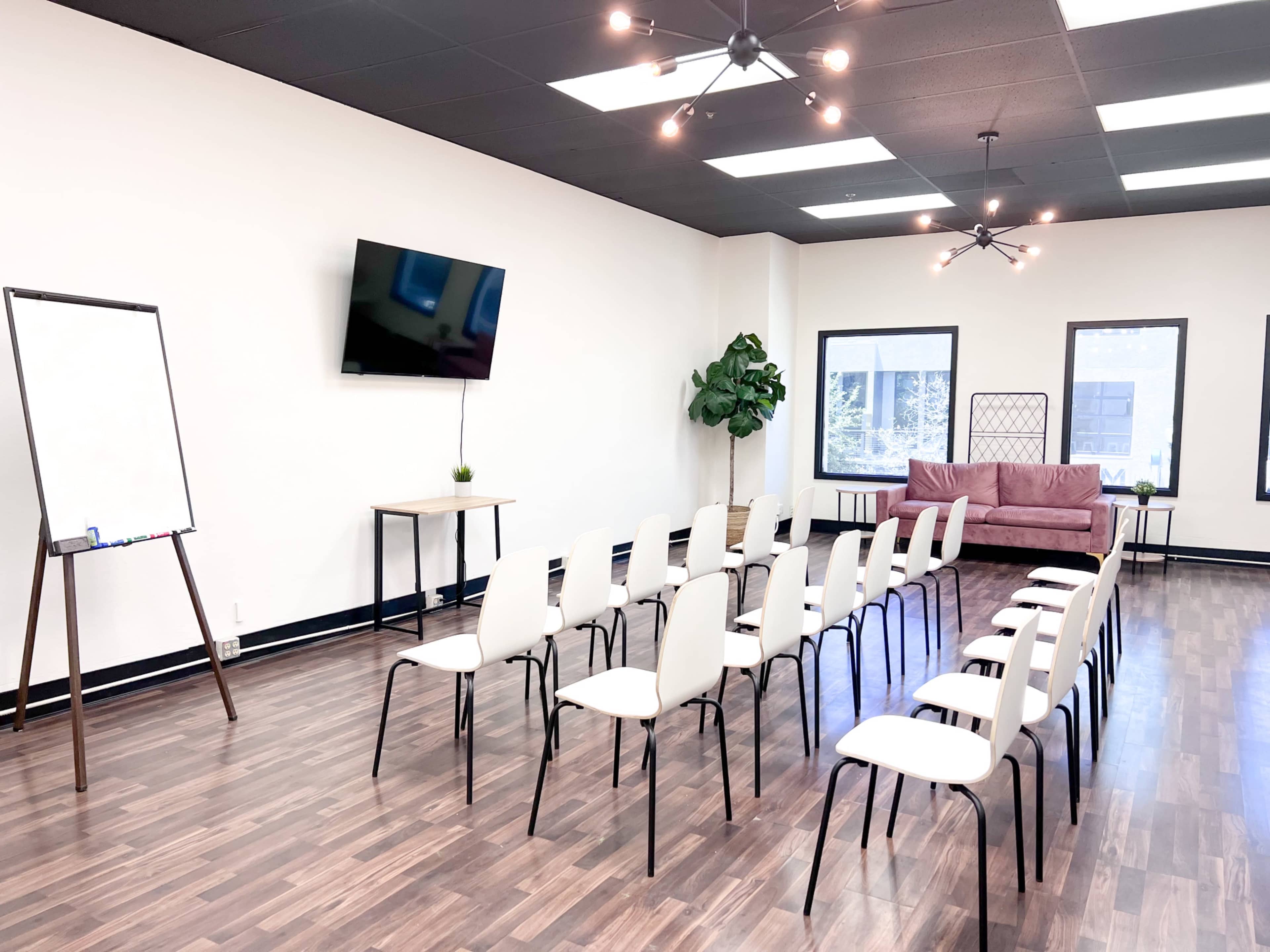 A conference room with a row of white chairs arranged facing a blank easel, a television mounted on the wall, and a couch in the background.