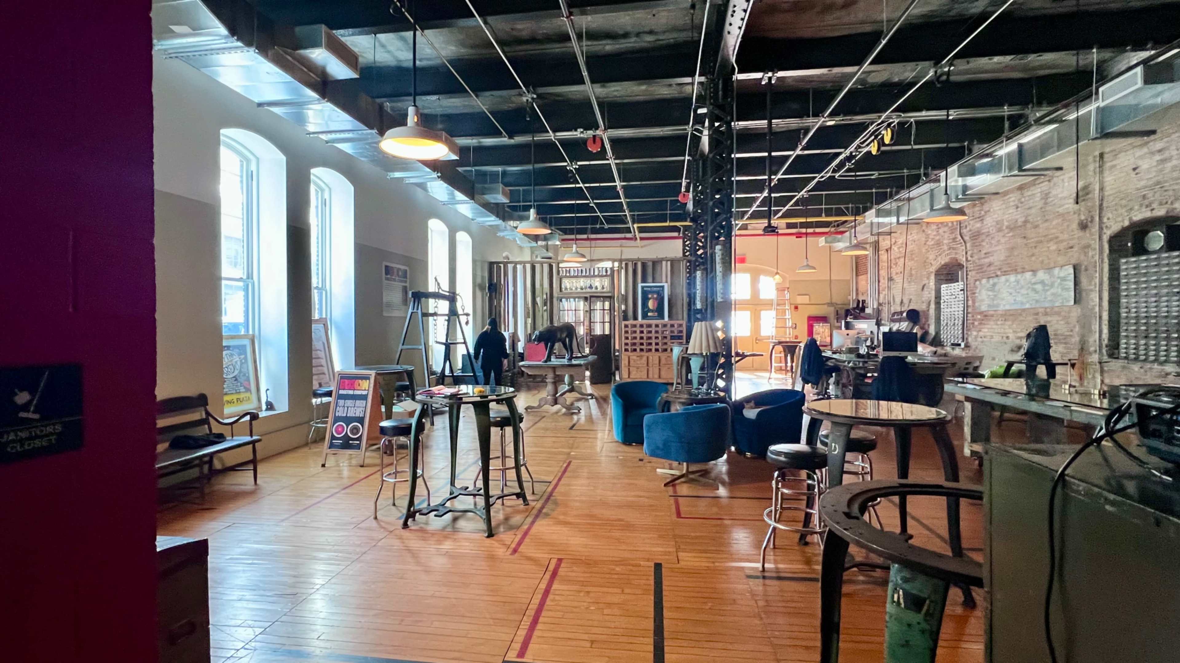 The image shows a spacious indoor venue with various seating arrangements, including tables and chairs, and people engaged in activity amidst exposed brick walls and industrial decor.