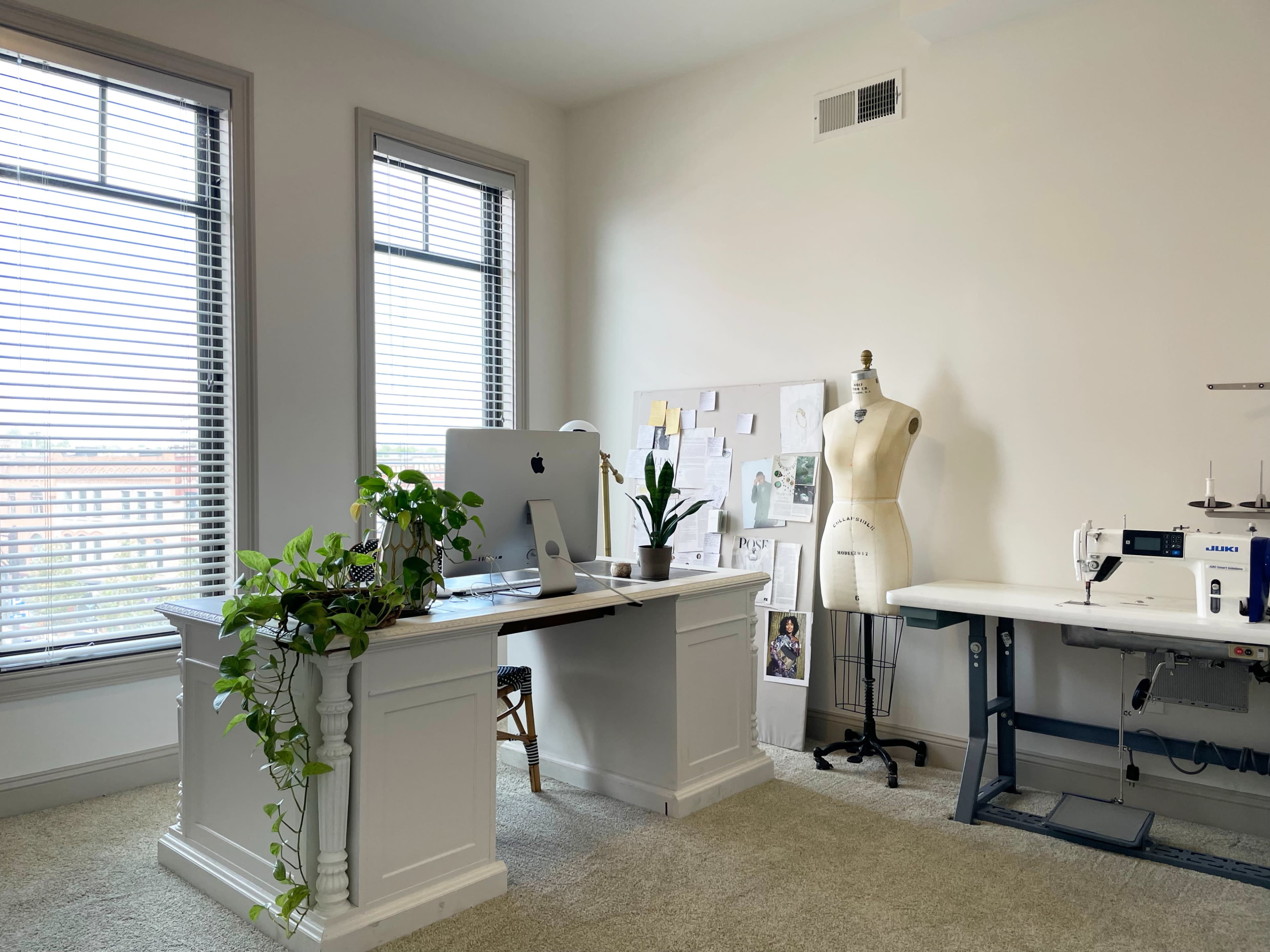 A bright office space features a desk with a computer, a sewing machine, and a dress form, alongside large windows and a plant on the desk.