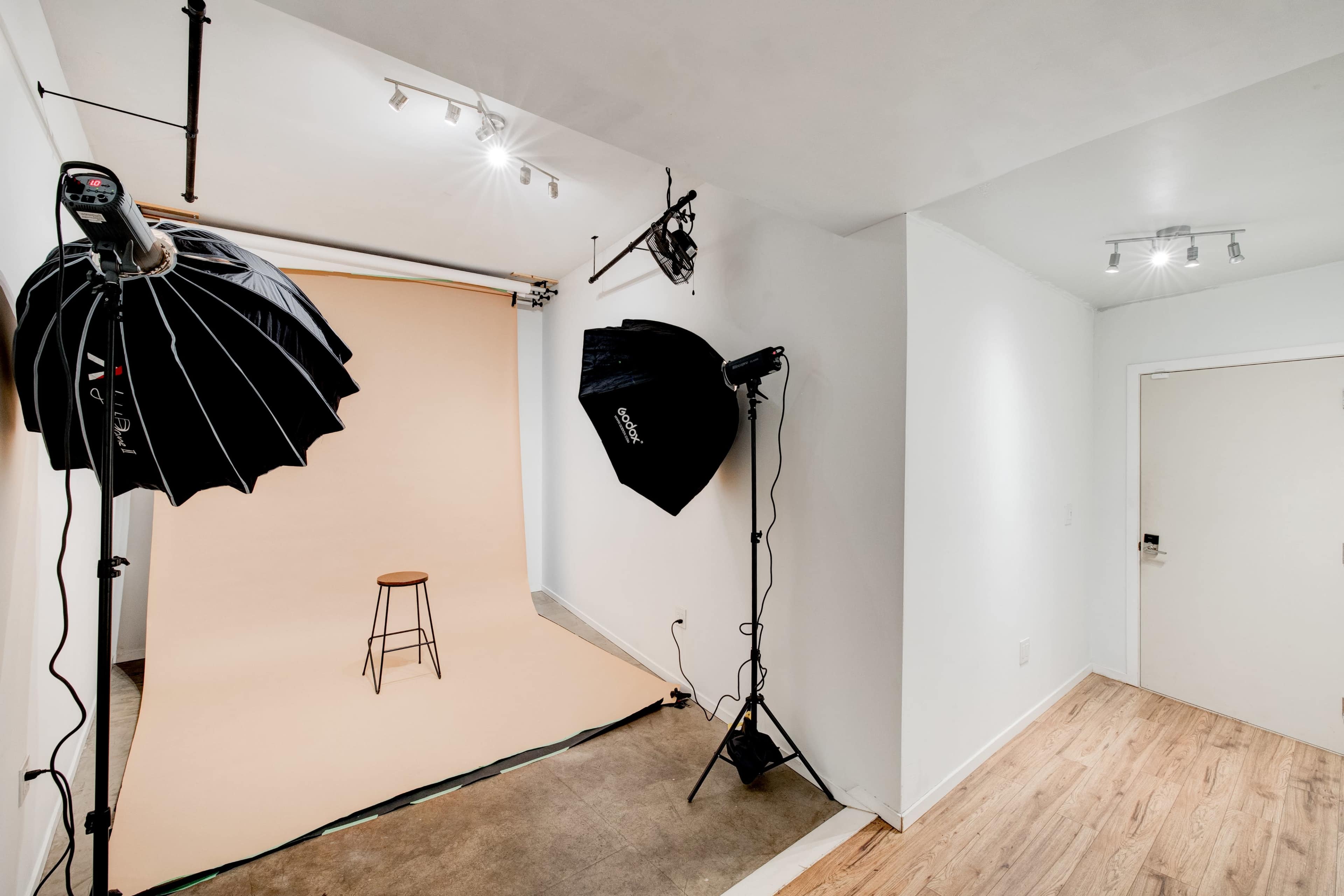 The image shows a photo studio setup with two large softbox lights, a beige backdrop, a wooden stool, and a doorway in a well-lit room.