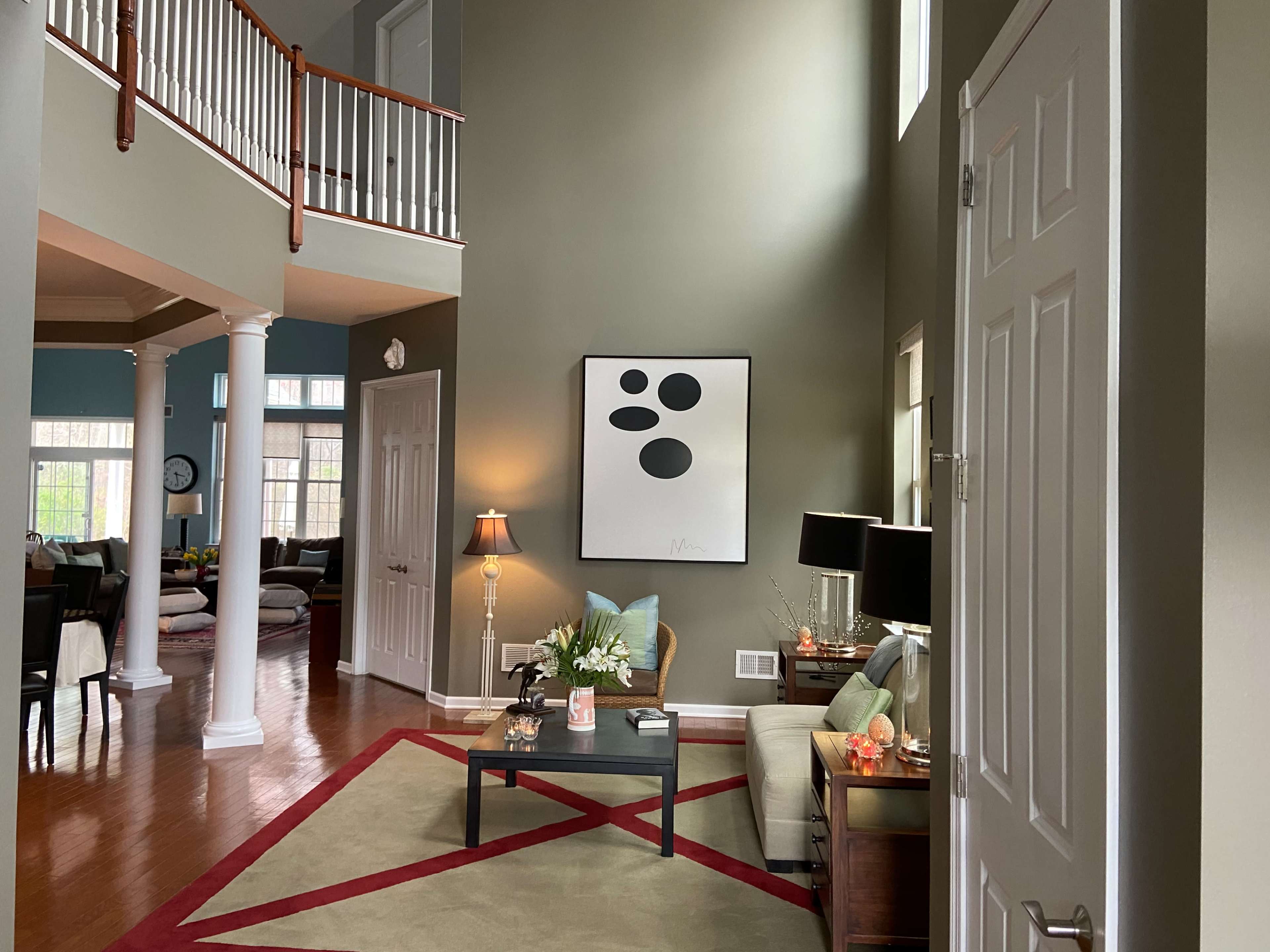 A spacious, well-lit living room featuring a high ceiling, contemporary artwork on the wall, and a mix of furniture arrangements on a hardwood floor.