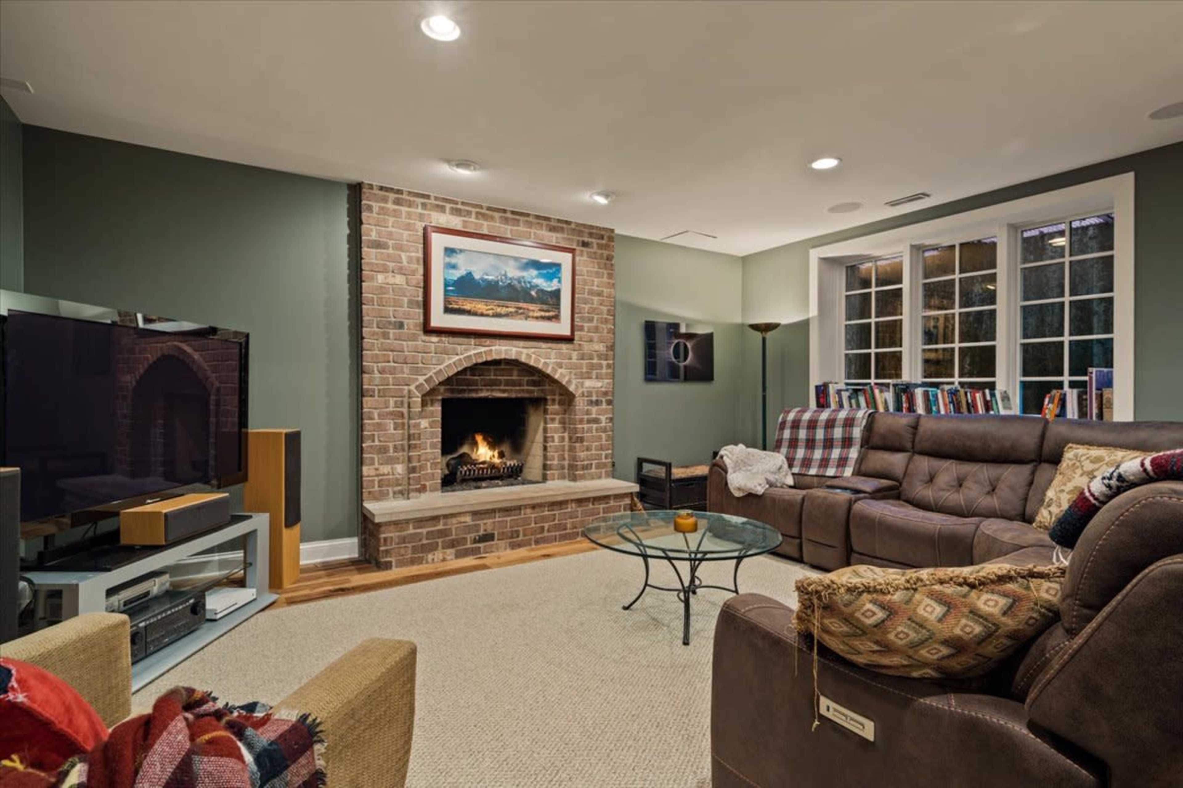 A cozy living room features a brick fireplace, a large sectional couch, a glass coffee table, and two televisions mounted on the walls.