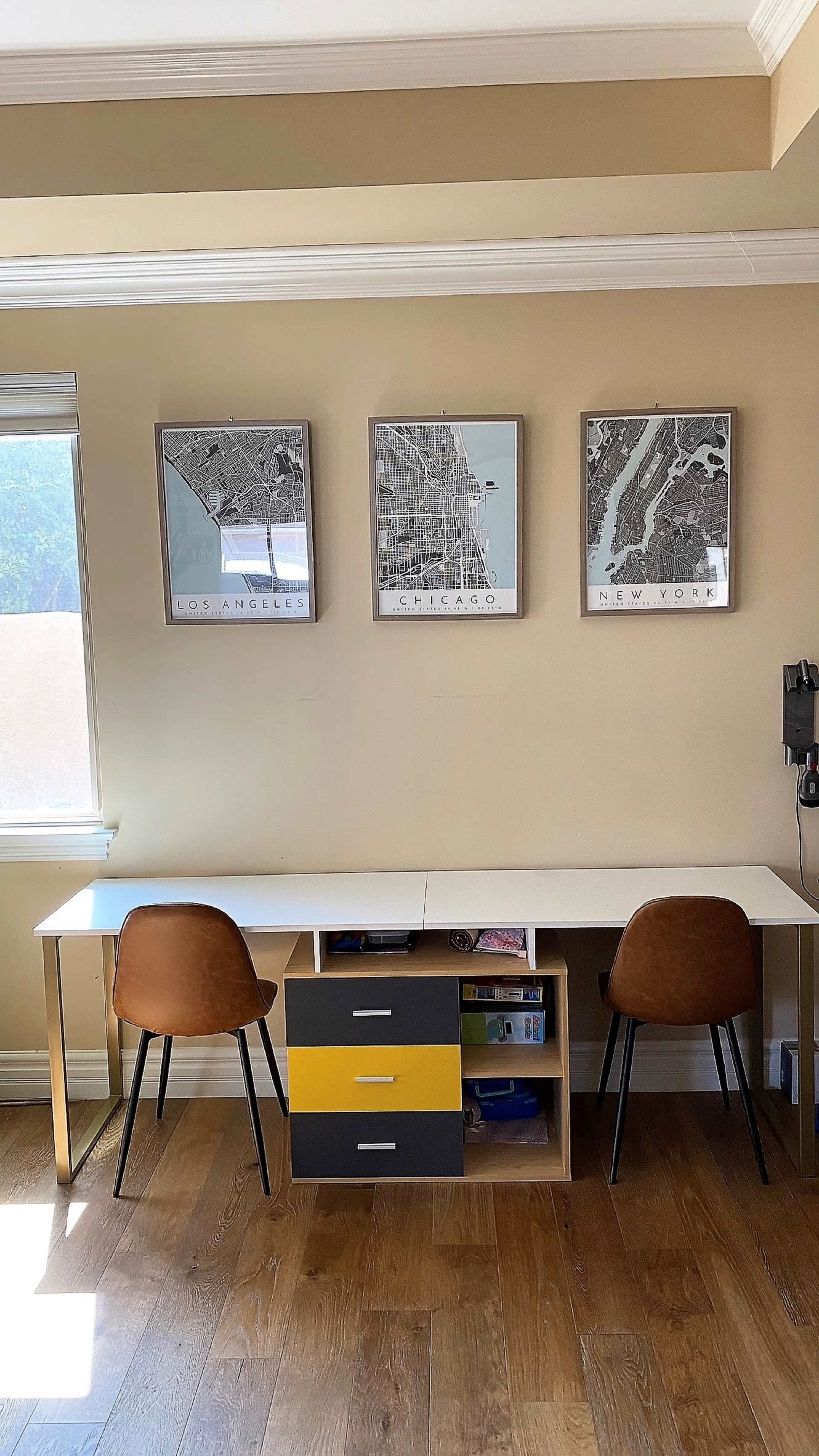 A minimalist workspace with a long white desk, two brown chairs, and a storage unit with yellow, gray, and white drawers, framed city map posters of Los Angeles, Chicago, and New York hanging on the wall.