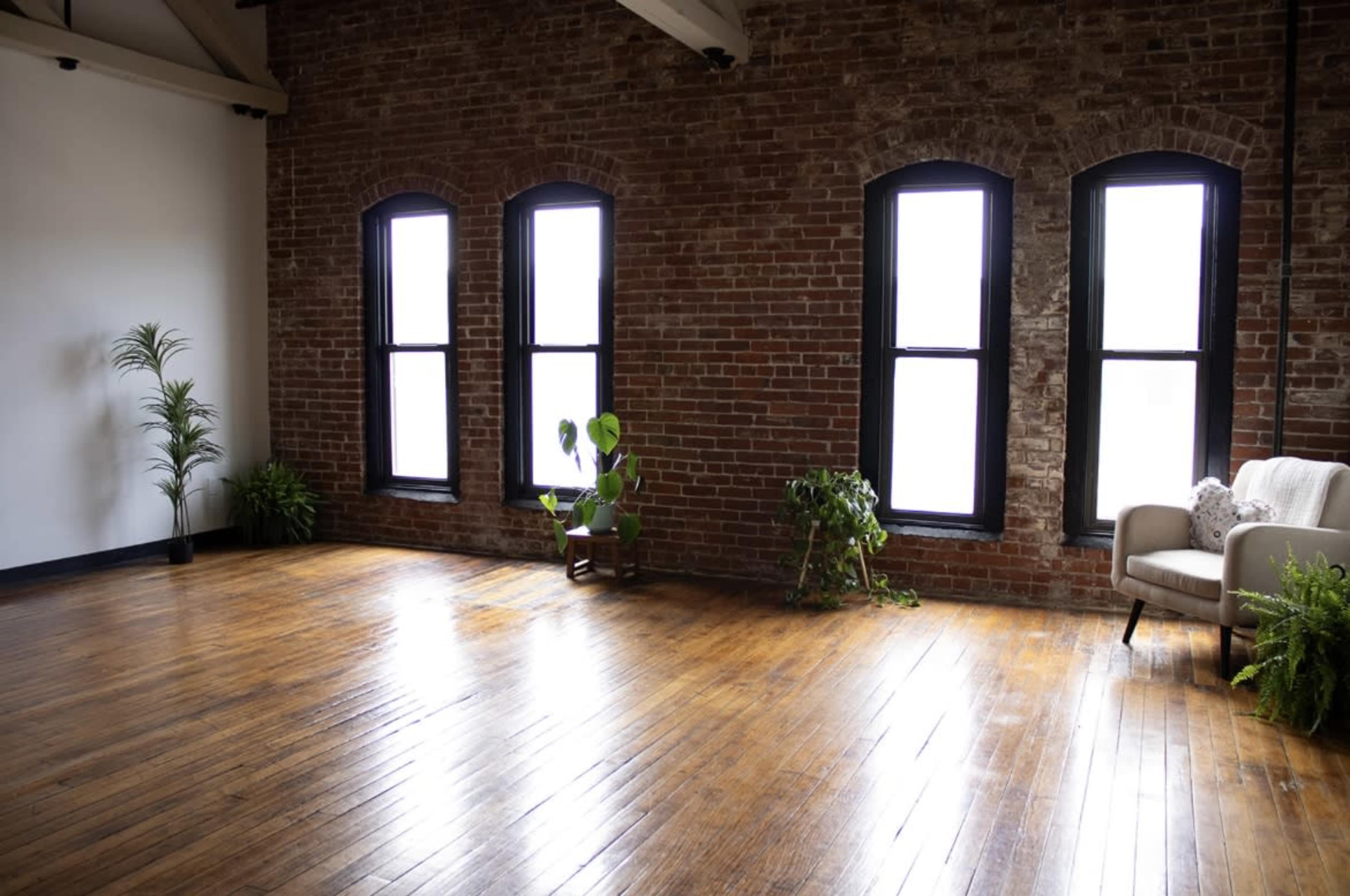 The image shows an empty room with exposed brick walls, wooden flooring, and large windows, featuring a few indoor plants and a single beige armchair.
