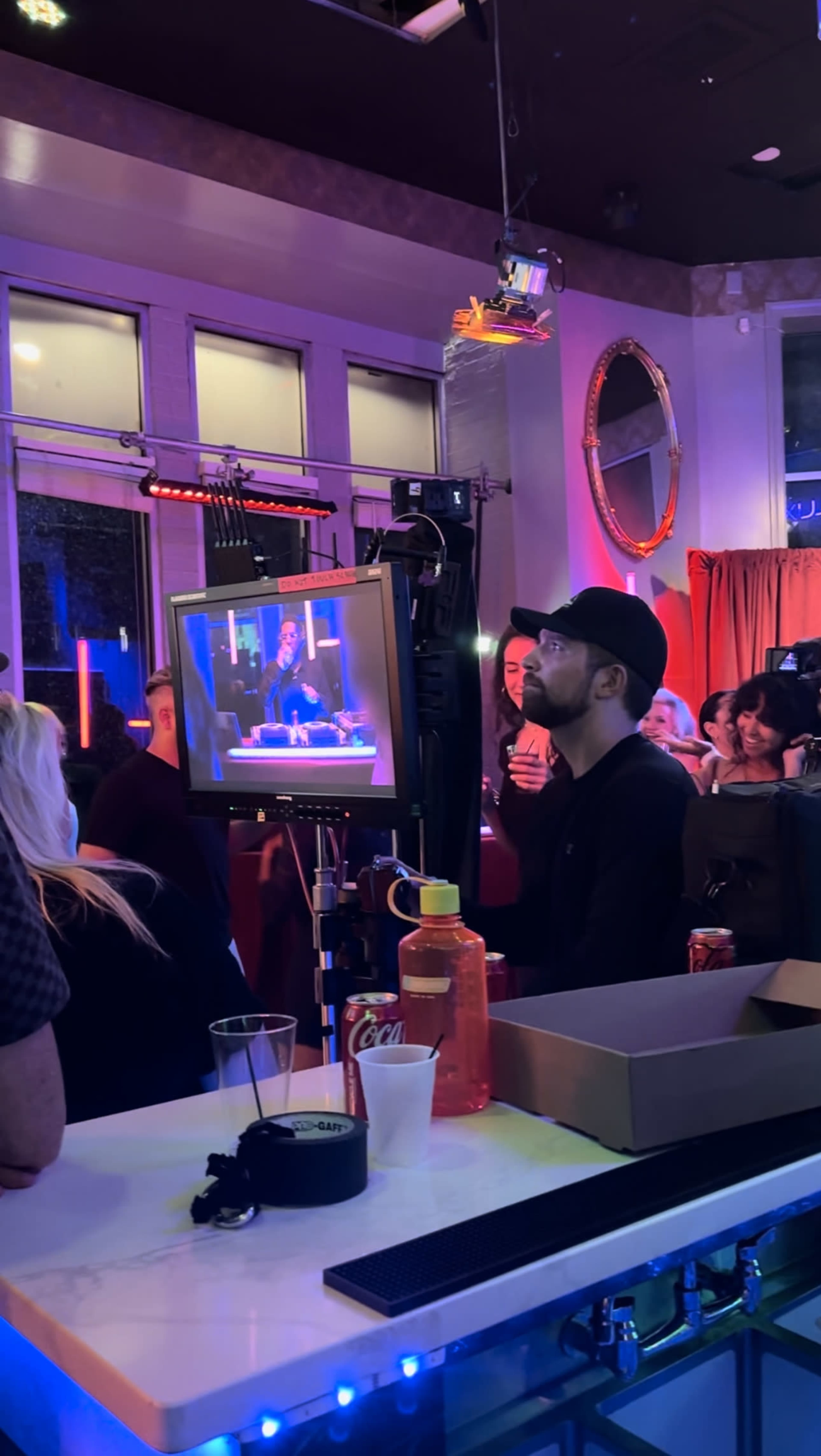 A man in a cap operates a camera setup in a vibrant bar setting, capturing a performance as patrons gather around.