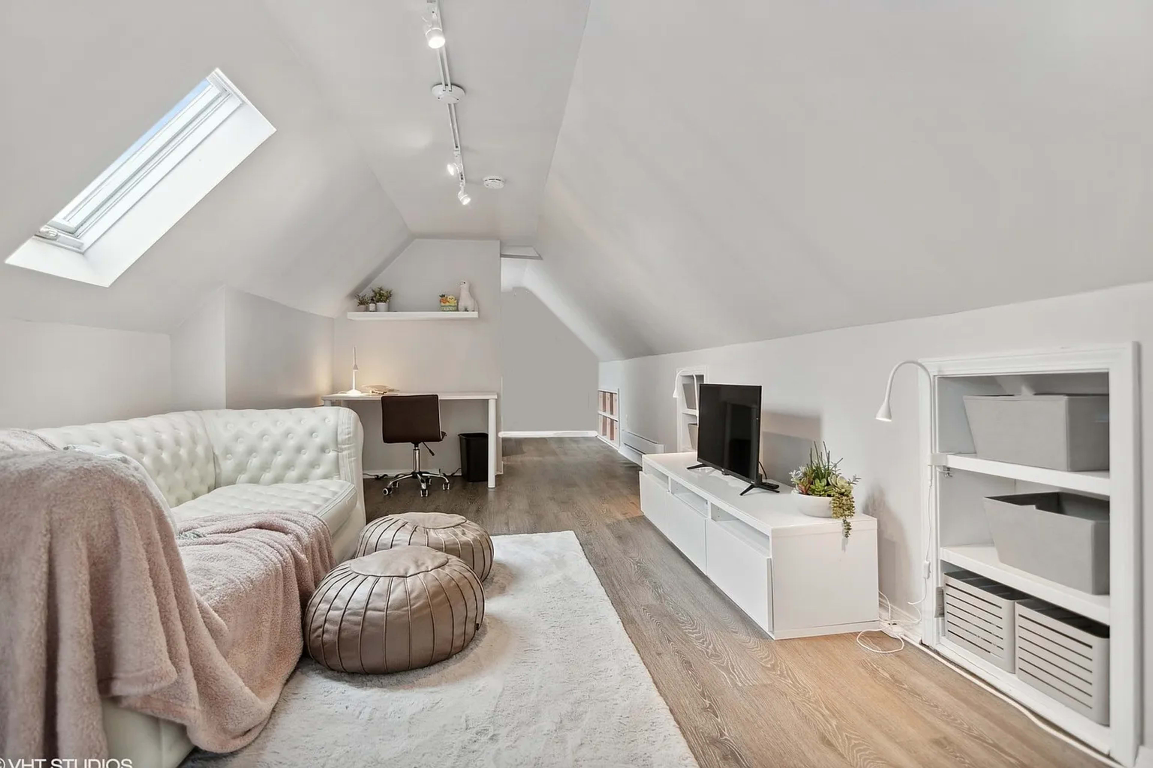 A modern attic space with a white sofa, a desk with a chair, a TV stand, and two large cushioned poufs.