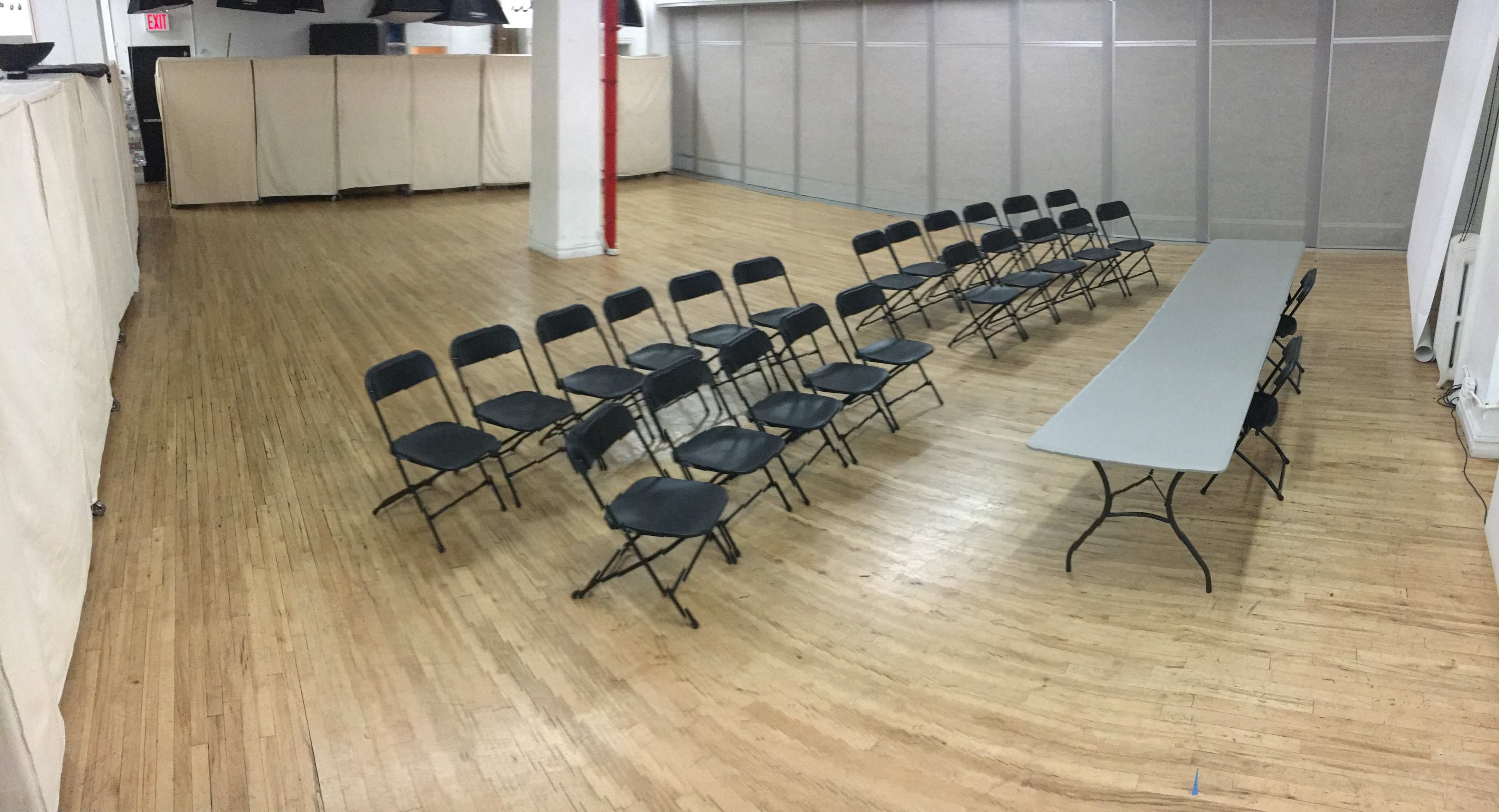 A spacious room with wooden flooring arranged with several folding chairs facing a long rectangular table.