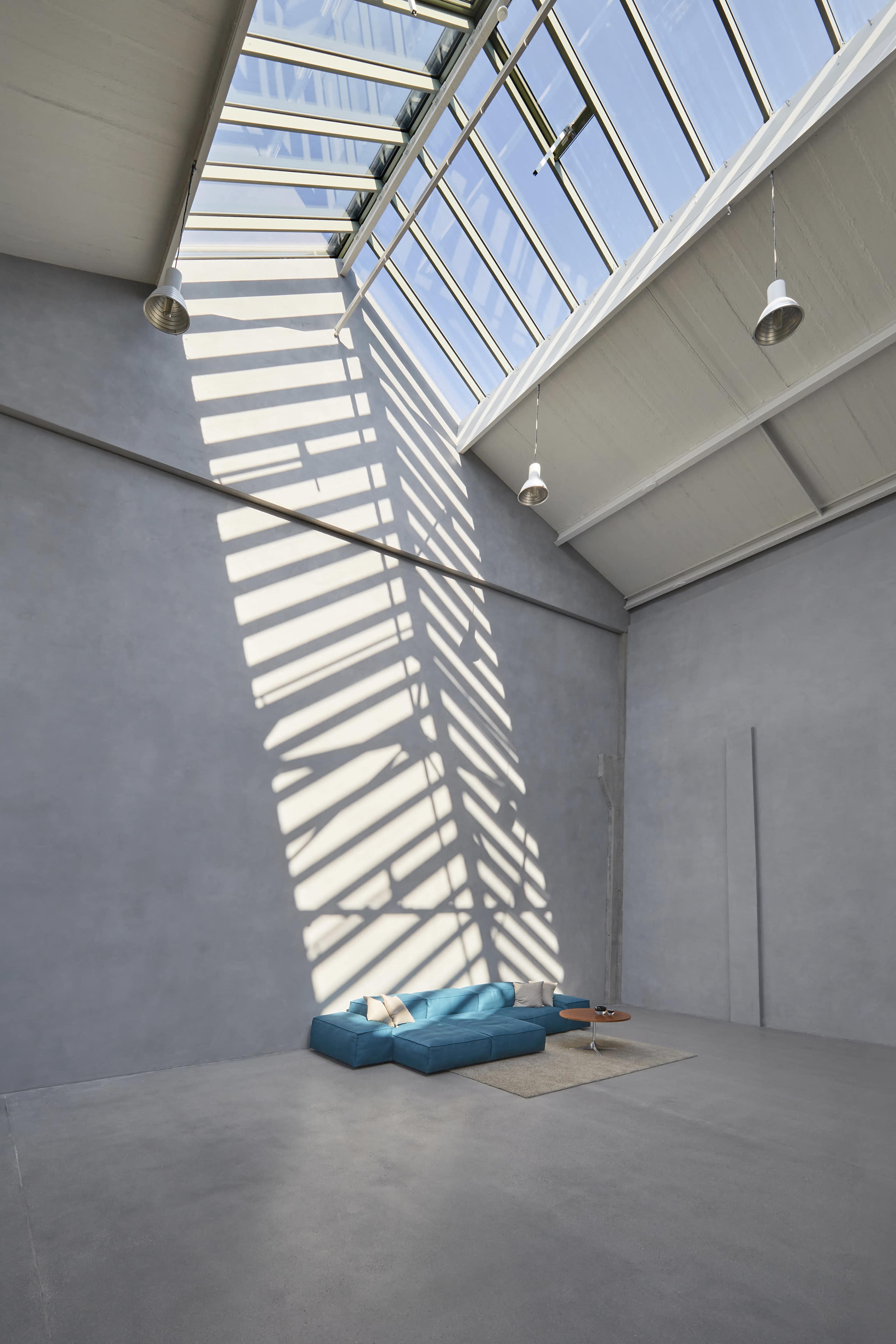 A modern interior space features a blue lounging area with a rug, illuminated by sunlight streaming through a skylight overhead.