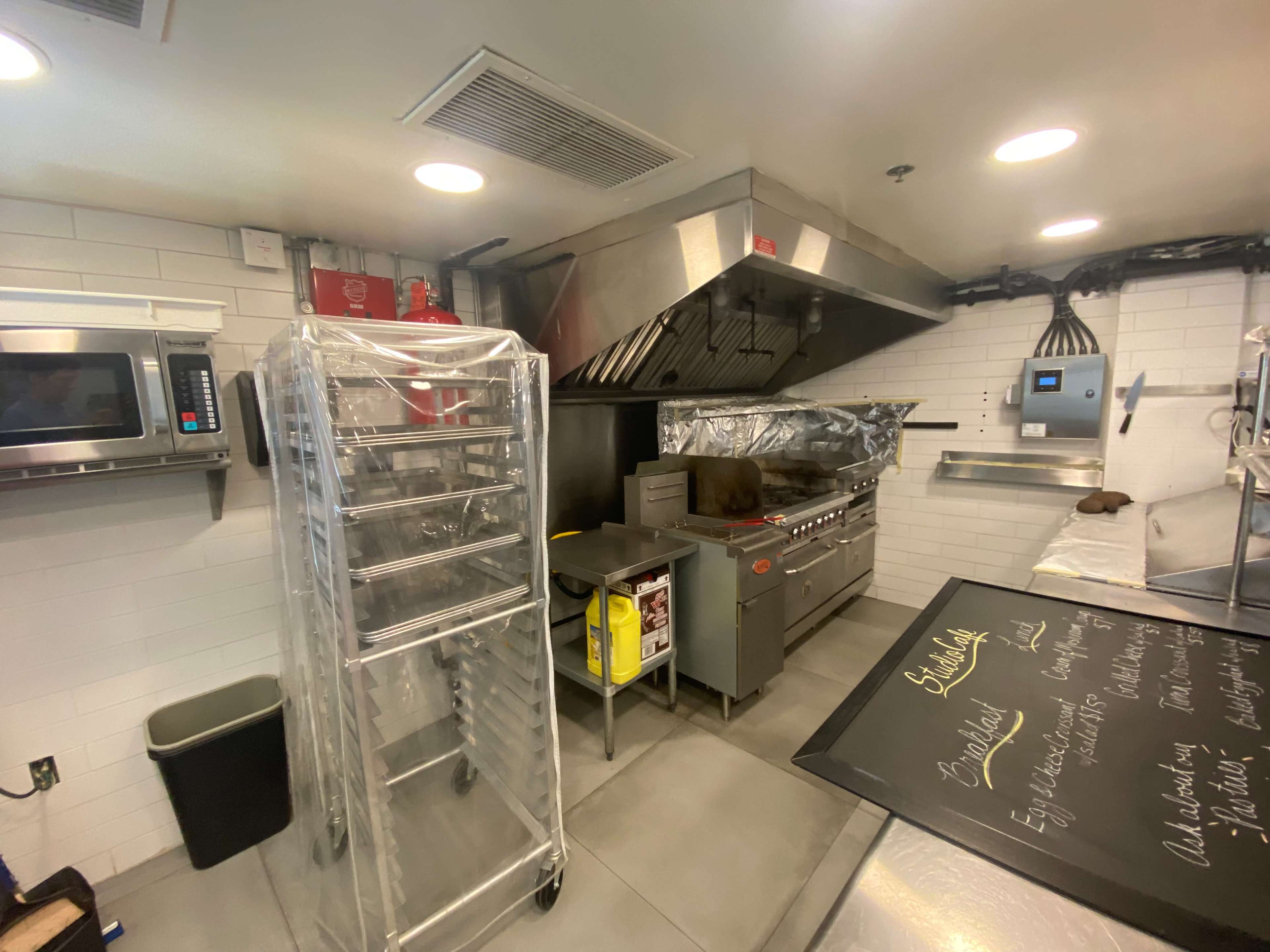 A commercial kitchen with stainless steel appliances, including a gas range, microwave, and a clear plastic-covered rack.