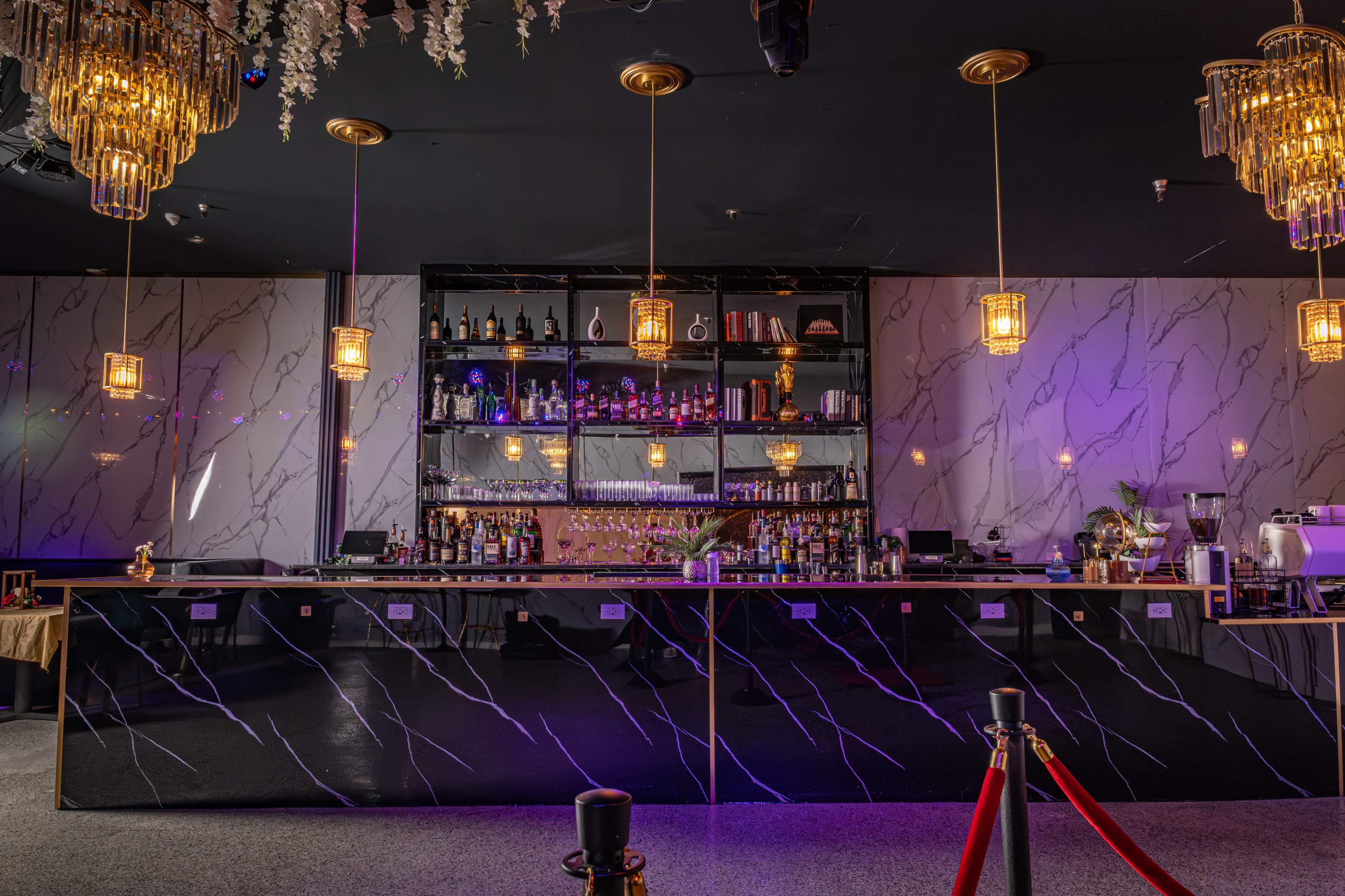 The image shows a stylish bar interior featuring a black marble bar counter, shelves displaying various bottles, and elegant chandeliers.