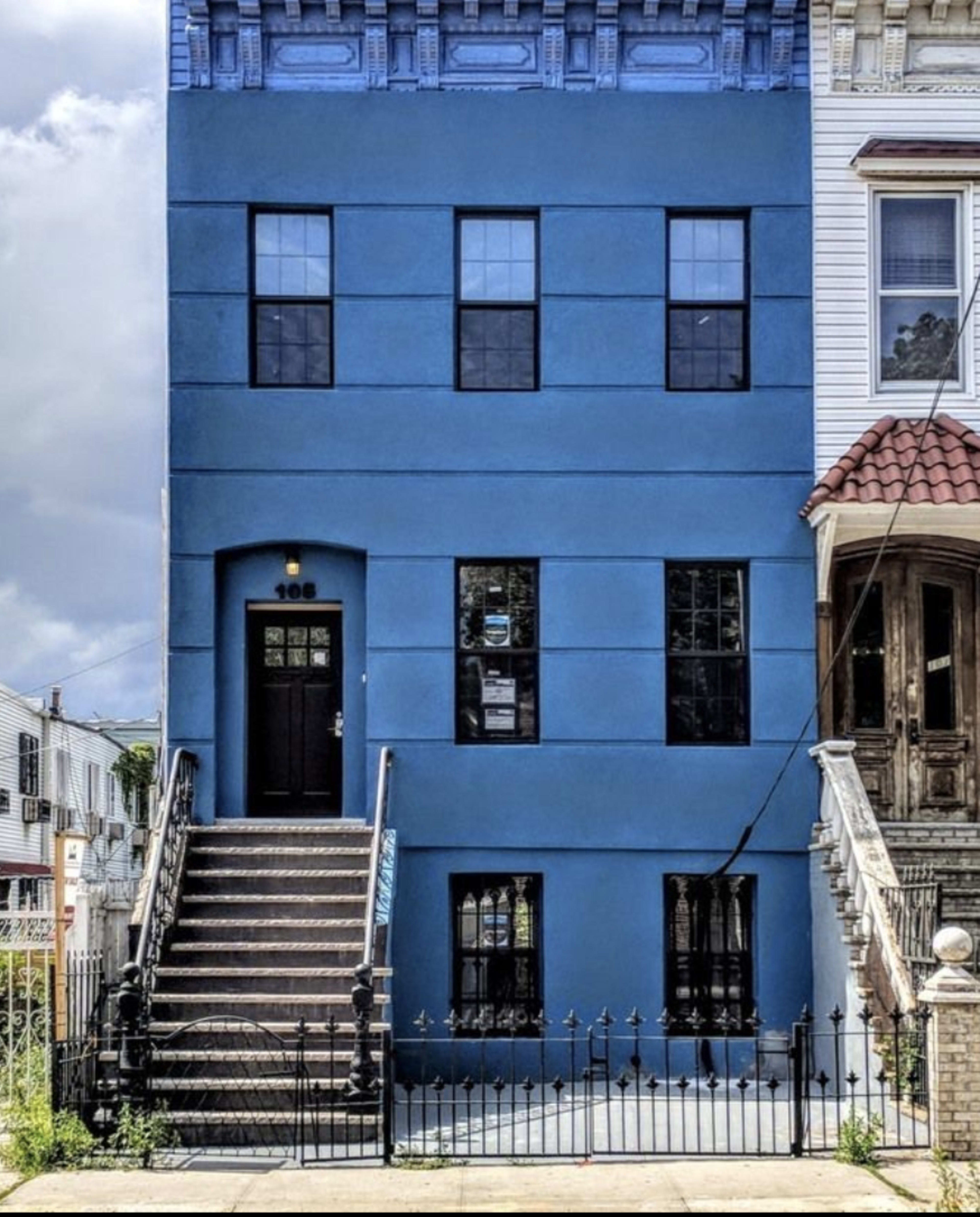 A blue three-story building with black windows and a staircase leading to the front door, located next to a building with a different architectural style.
