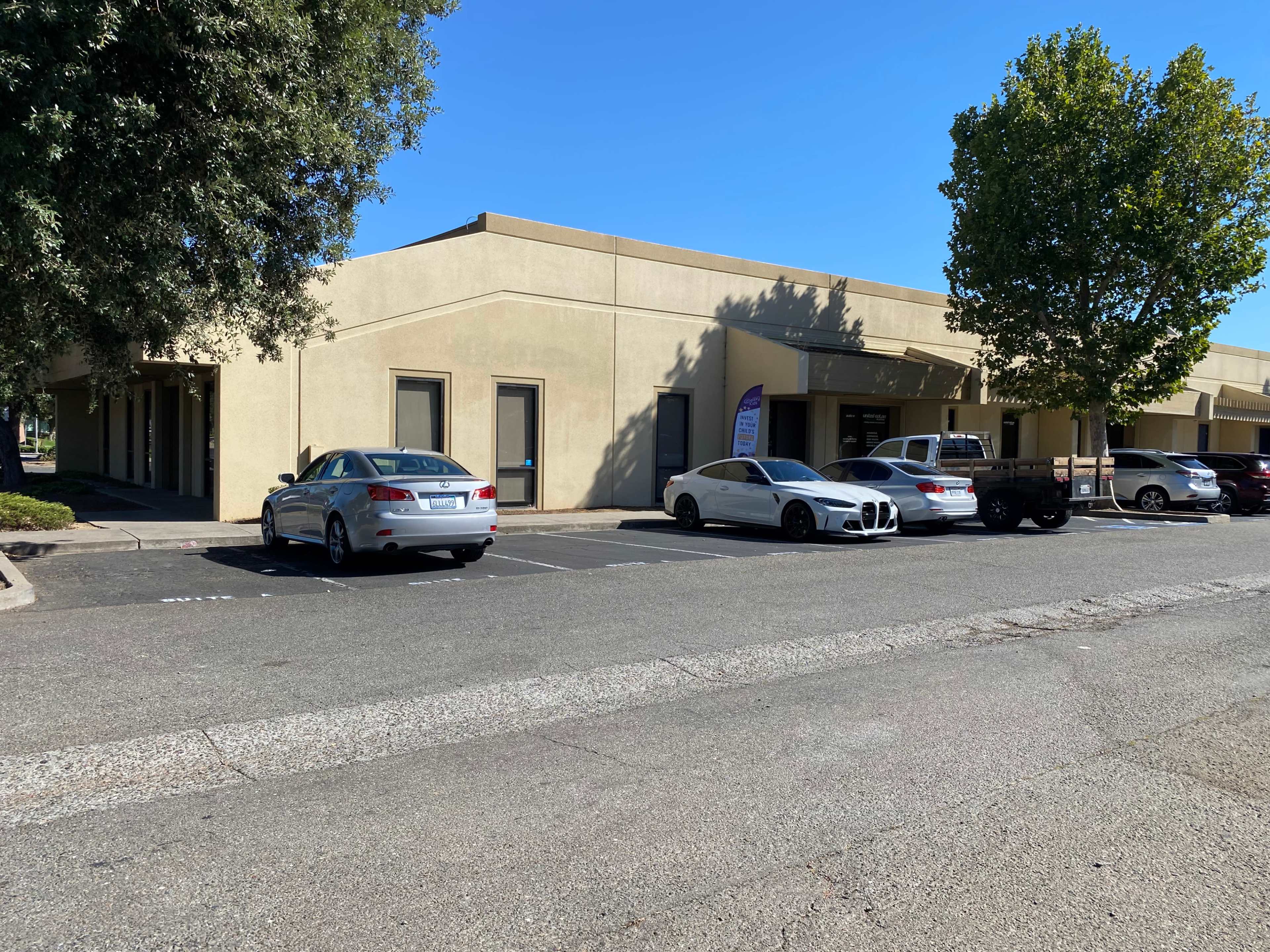 The image shows a row of commercial buildings with several parked cars in front of them under clear blue skies.