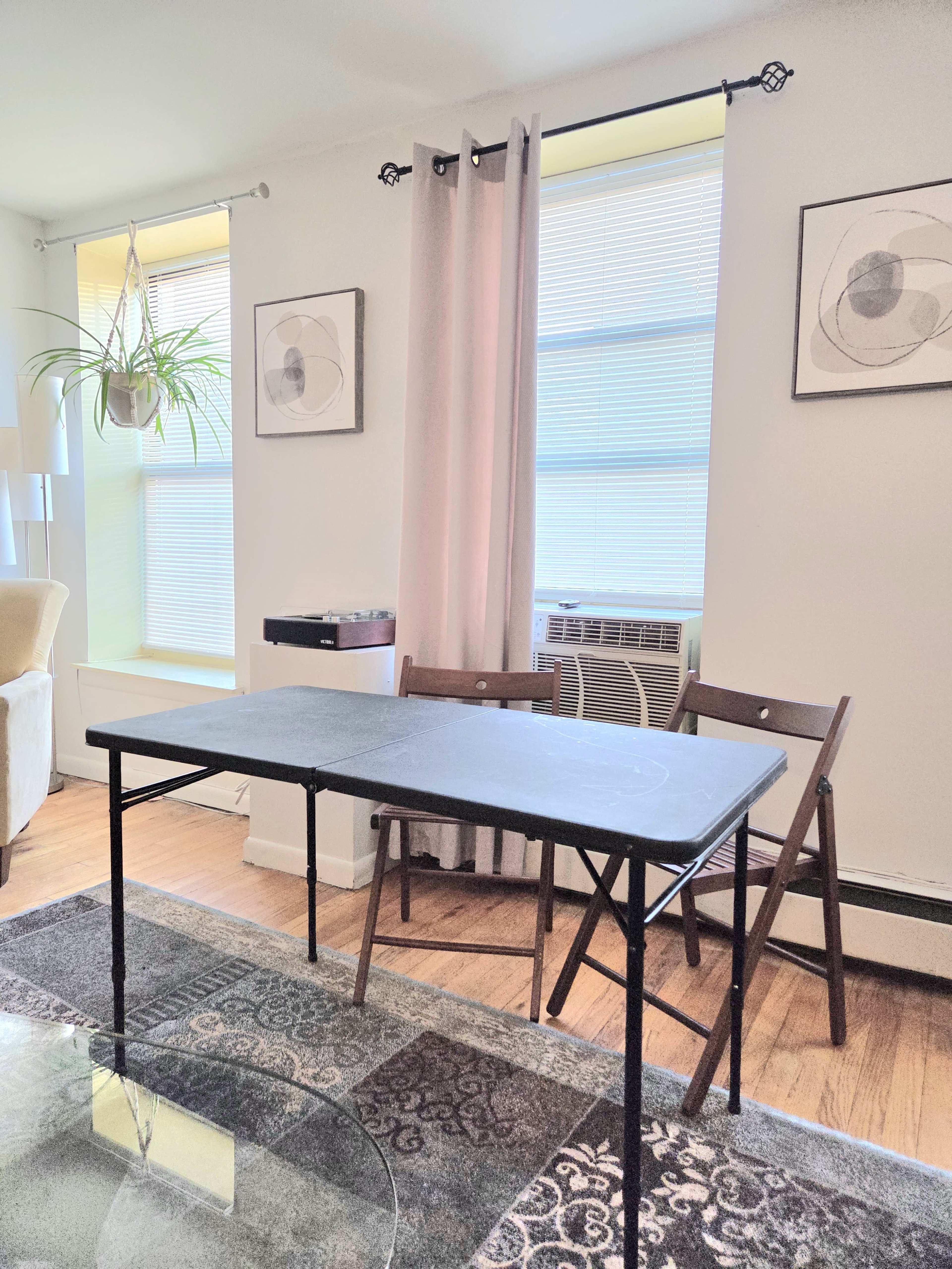 The image shows a small dining area featuring a black folding table with two wooden chairs, a rug on the floor, and large windows adorned with sheer curtains.
