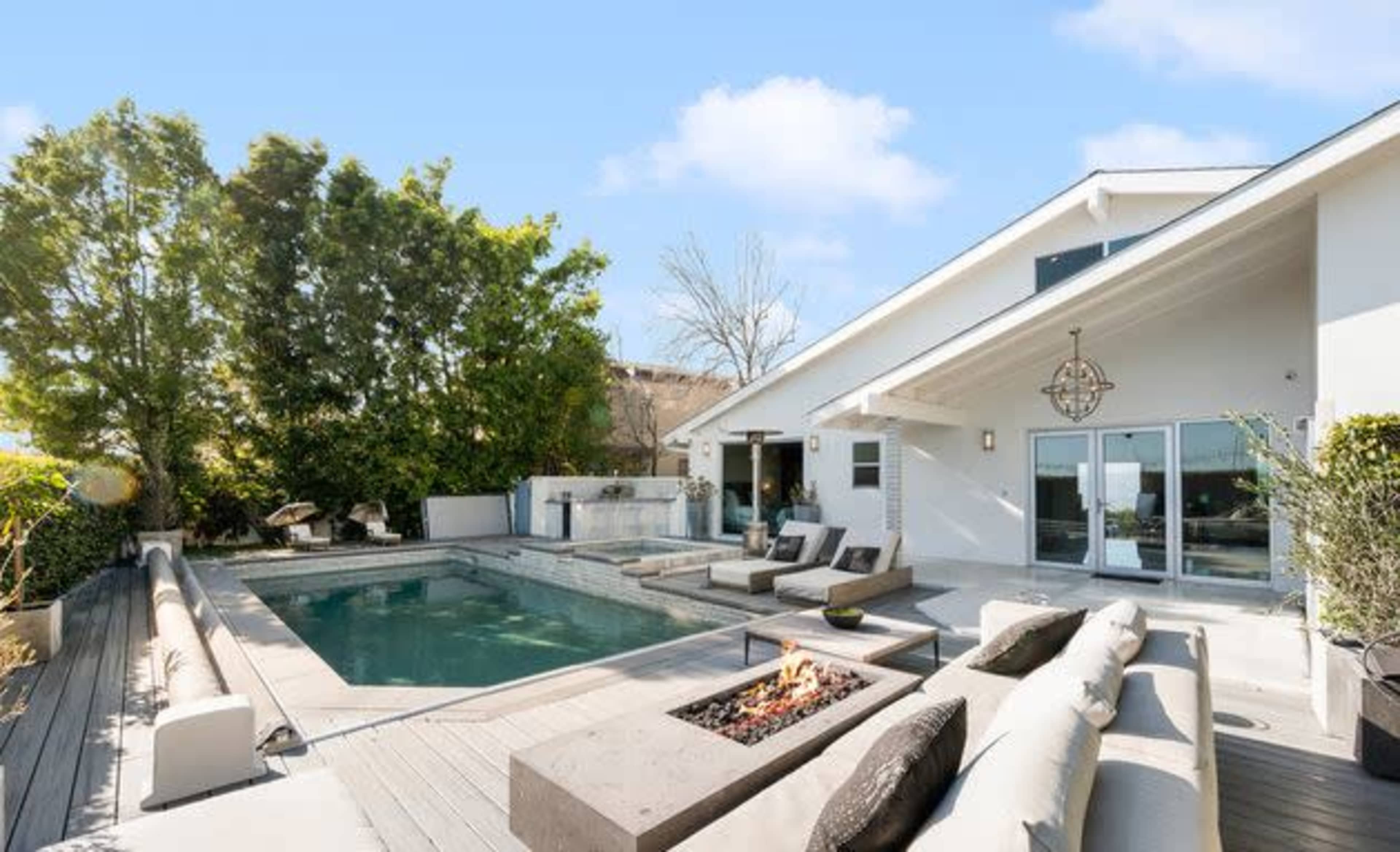 The image shows a modern outdoor area with a swimming pool, fire pit, and comfortable seating, surrounded by lush greenery and a house in the background.