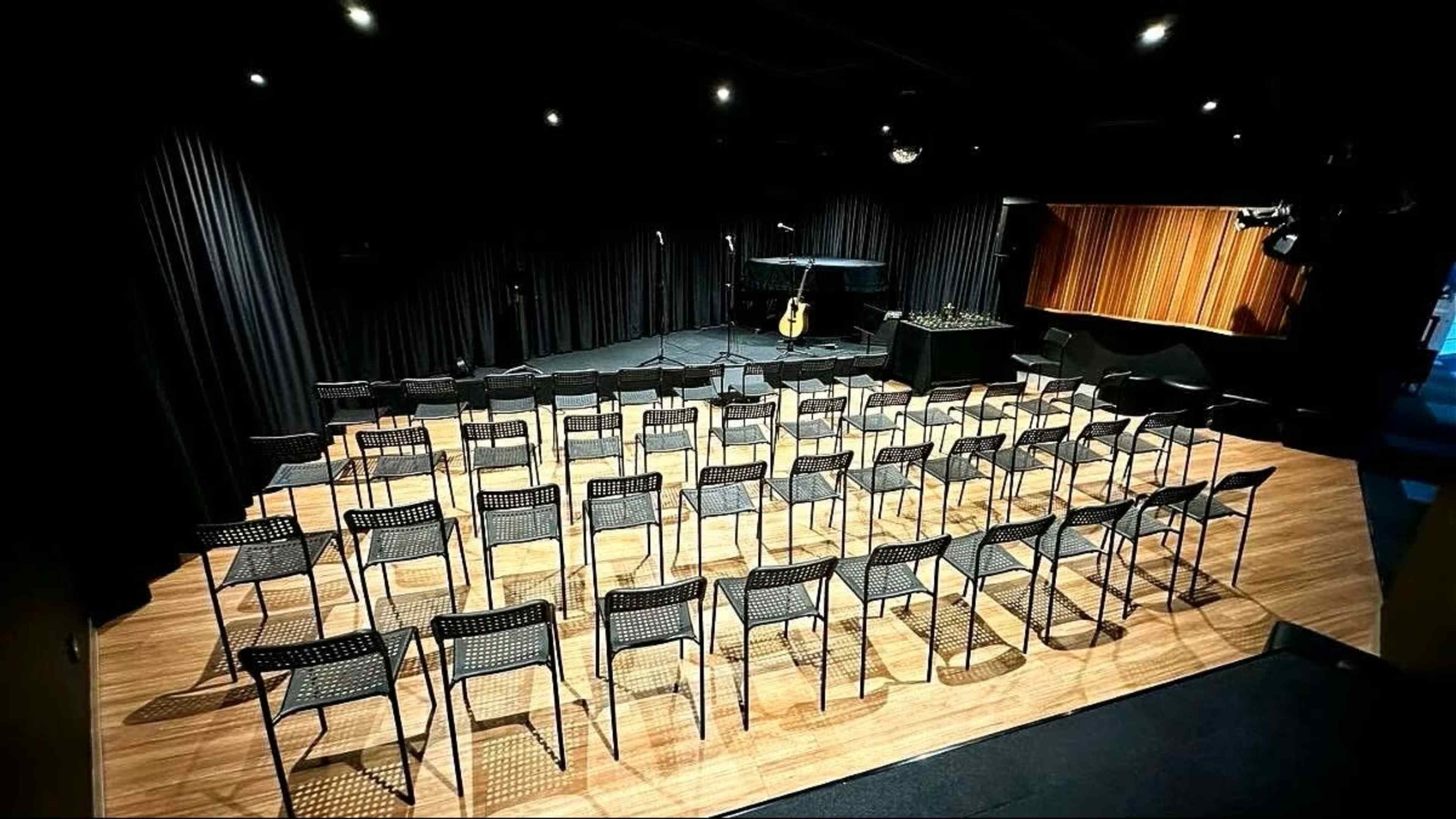 The image shows a music performance space with rows of empty chairs arranged on a wooden floor in front of a stage.