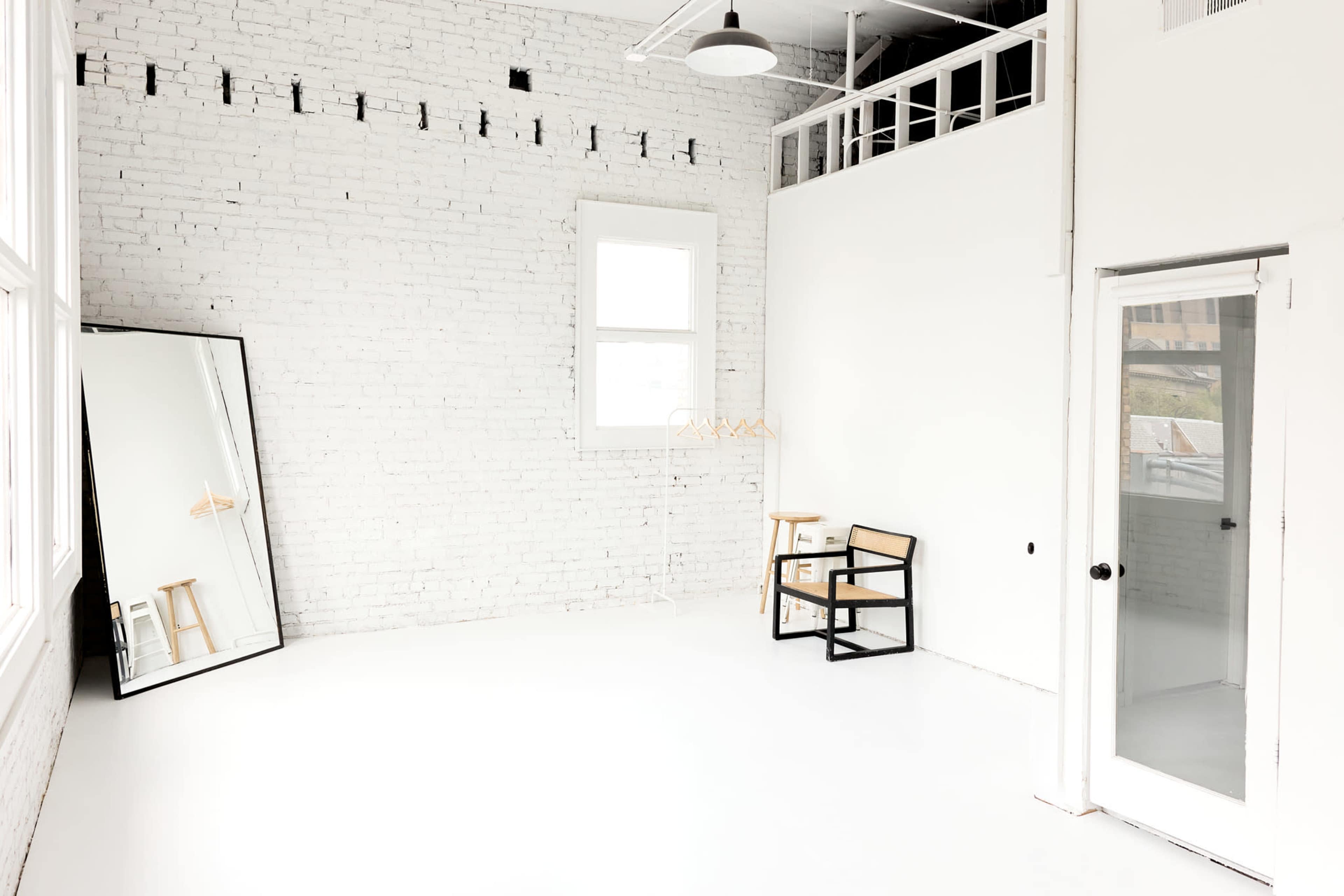 The image shows a bright, minimalist studio featuring white brick walls, a large mirror, two wooden chairs, and a glass door.