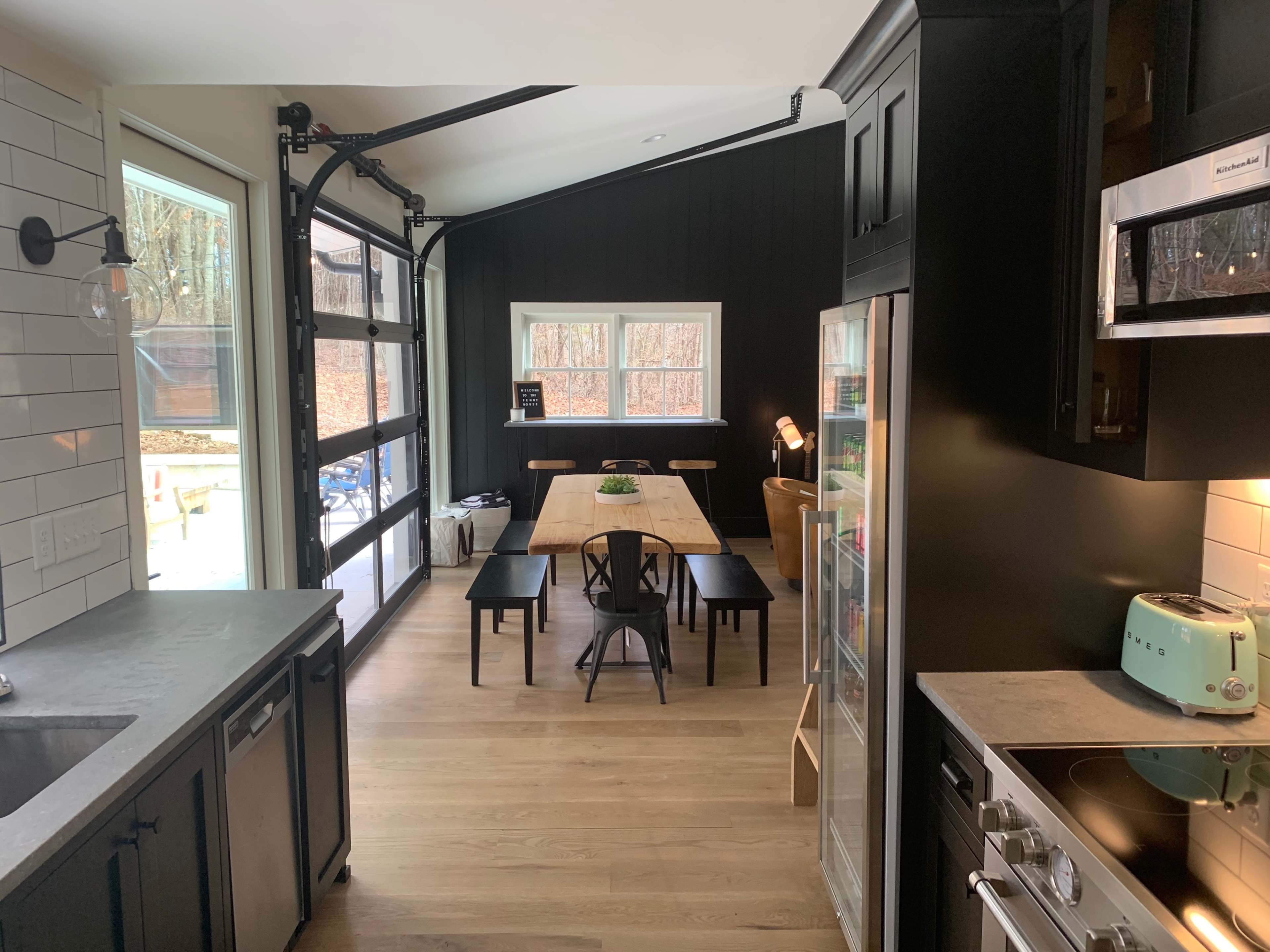 A modern kitchen with a view of a dining area featuring a long wooden table and large windows overlooking a backyard.