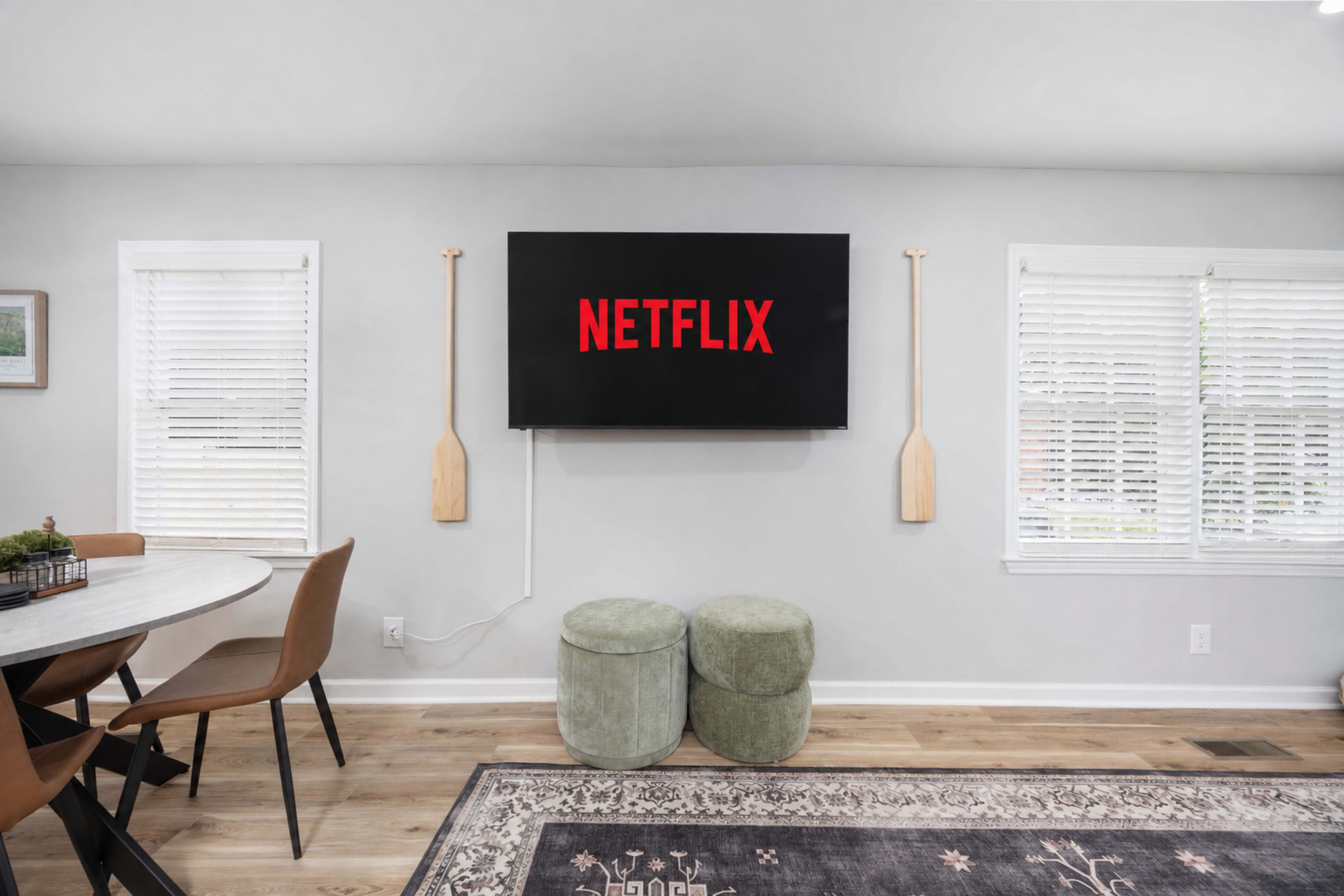 A television displaying the Netflix logo is mounted on a light gray wall, flanked by two wooden paddles, with a round dining table and green stools nearby.