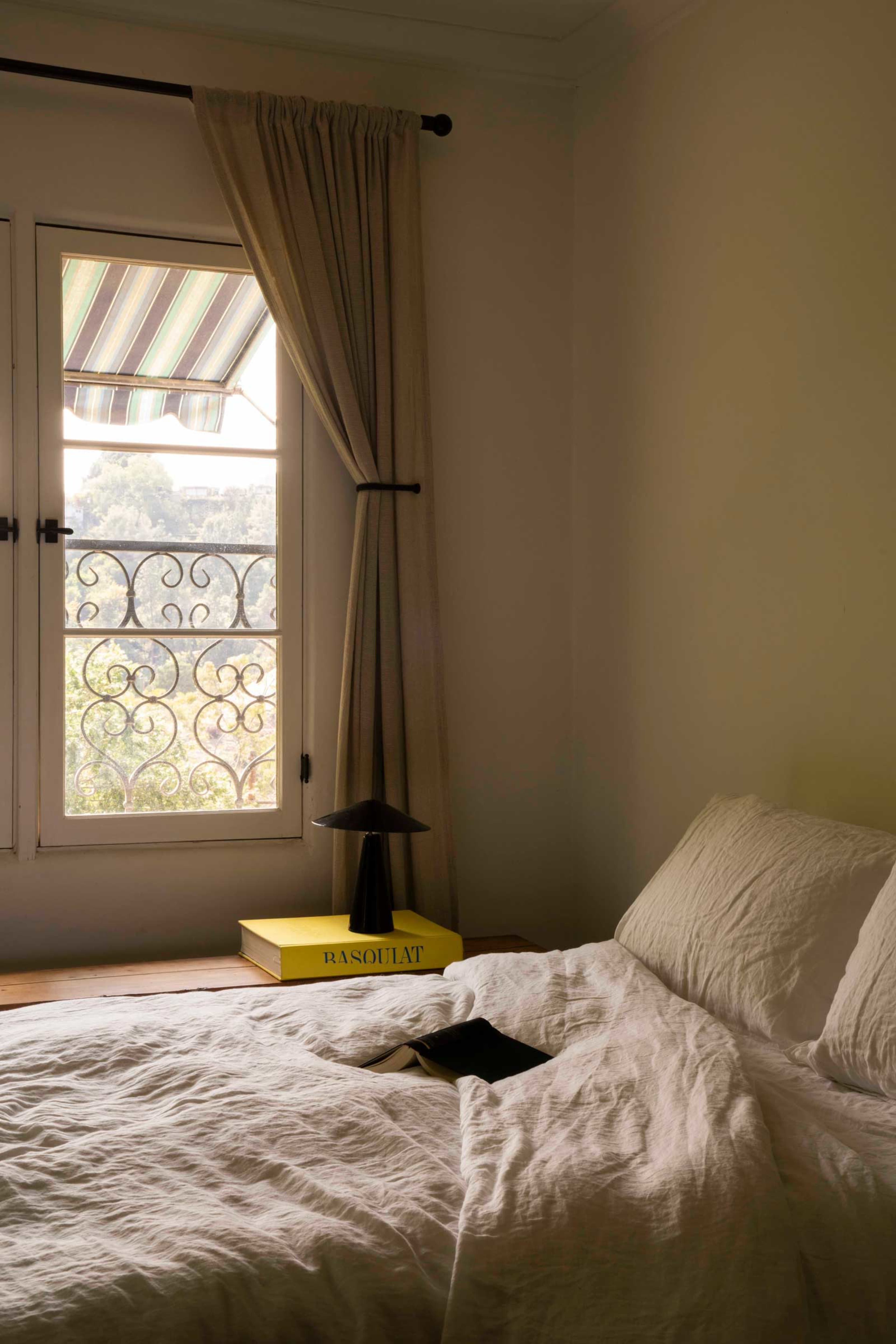 A neatly made bed with a wrinkled white duvet is positioned near a window featuring striped curtains and an iron railing.