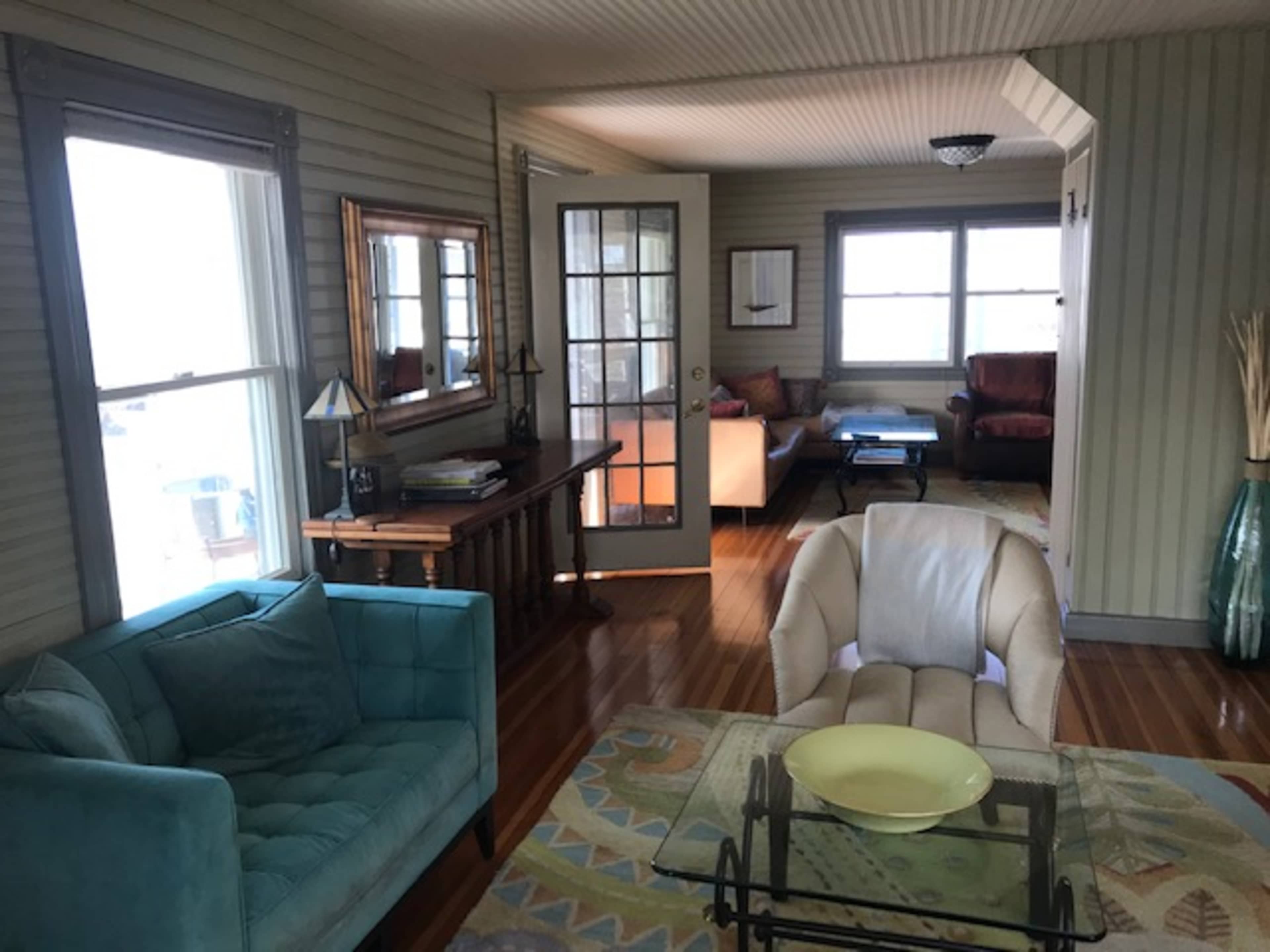 The scene depicts a cozy living area with a teal couch, a cream-colored armchair, a coffee table, and large windows allowing natural light.