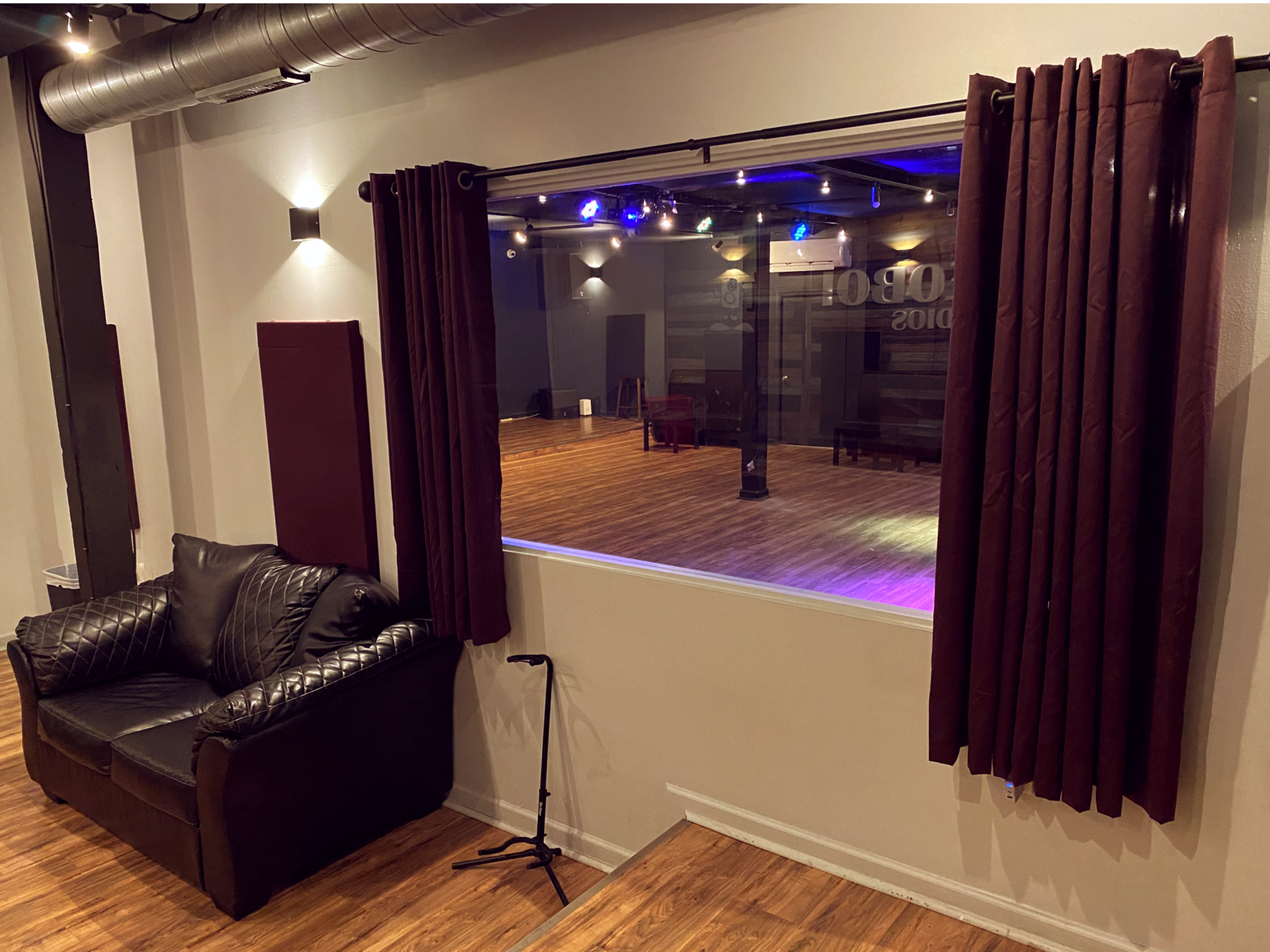 A cozy room with a black leather couch and a large window overlooking a wooden floor space with stage lights.
