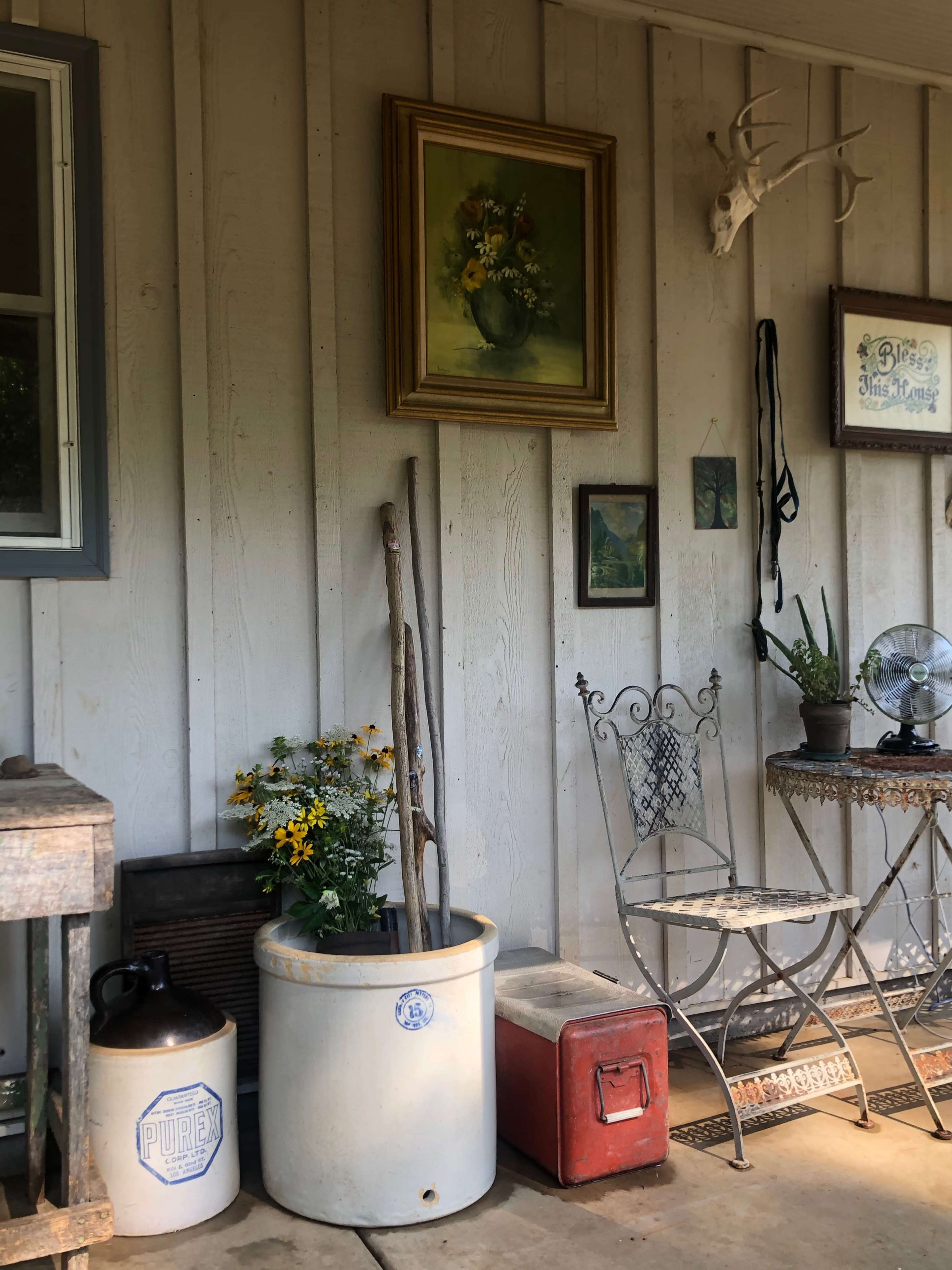 A rustic porch display features a wooden chair, a painted flower pot with sticks, an antique cooler, and framed artwork on the wall.