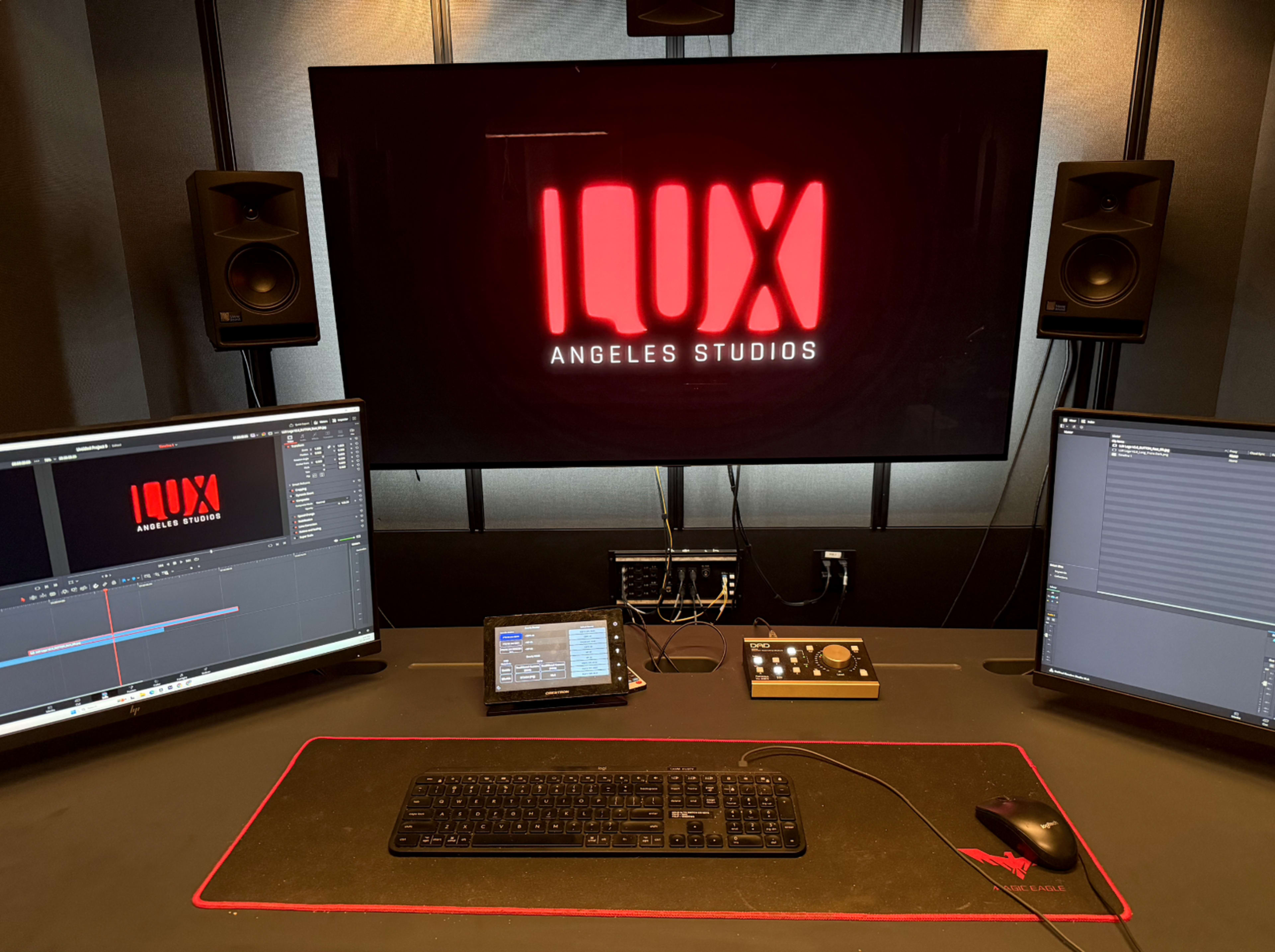 The image shows a modern audio-visual studio environment with a large backlit logo reading "ILUX ANGELES STUDIOS" on a screen, alongside multiple monitors and a control interface on a desk.