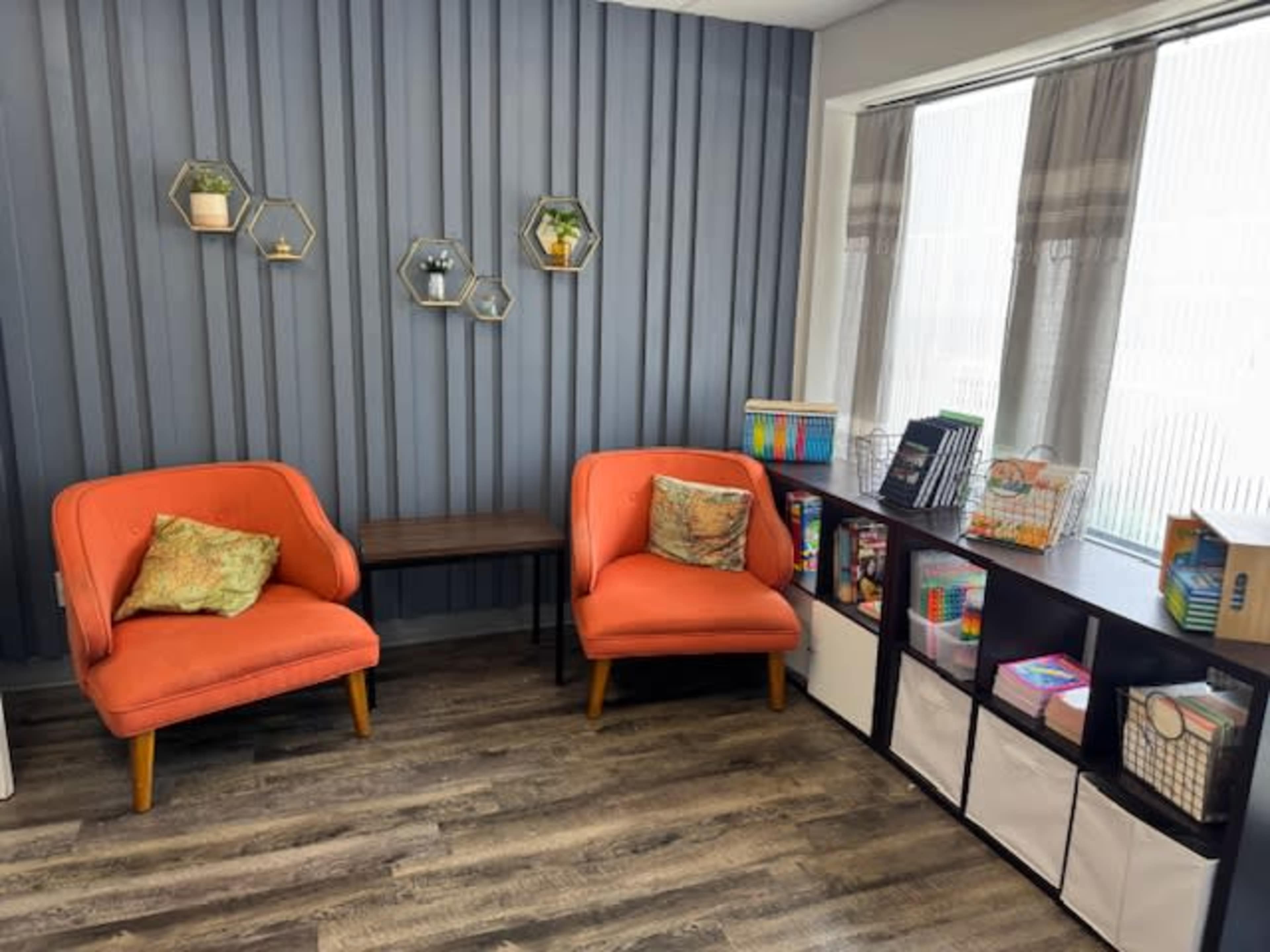 The image shows a small, well-lit room with two orange armchairs, a wooden table, and a shelving unit filled with various colorful items and organizational bins against a striped blue wall.