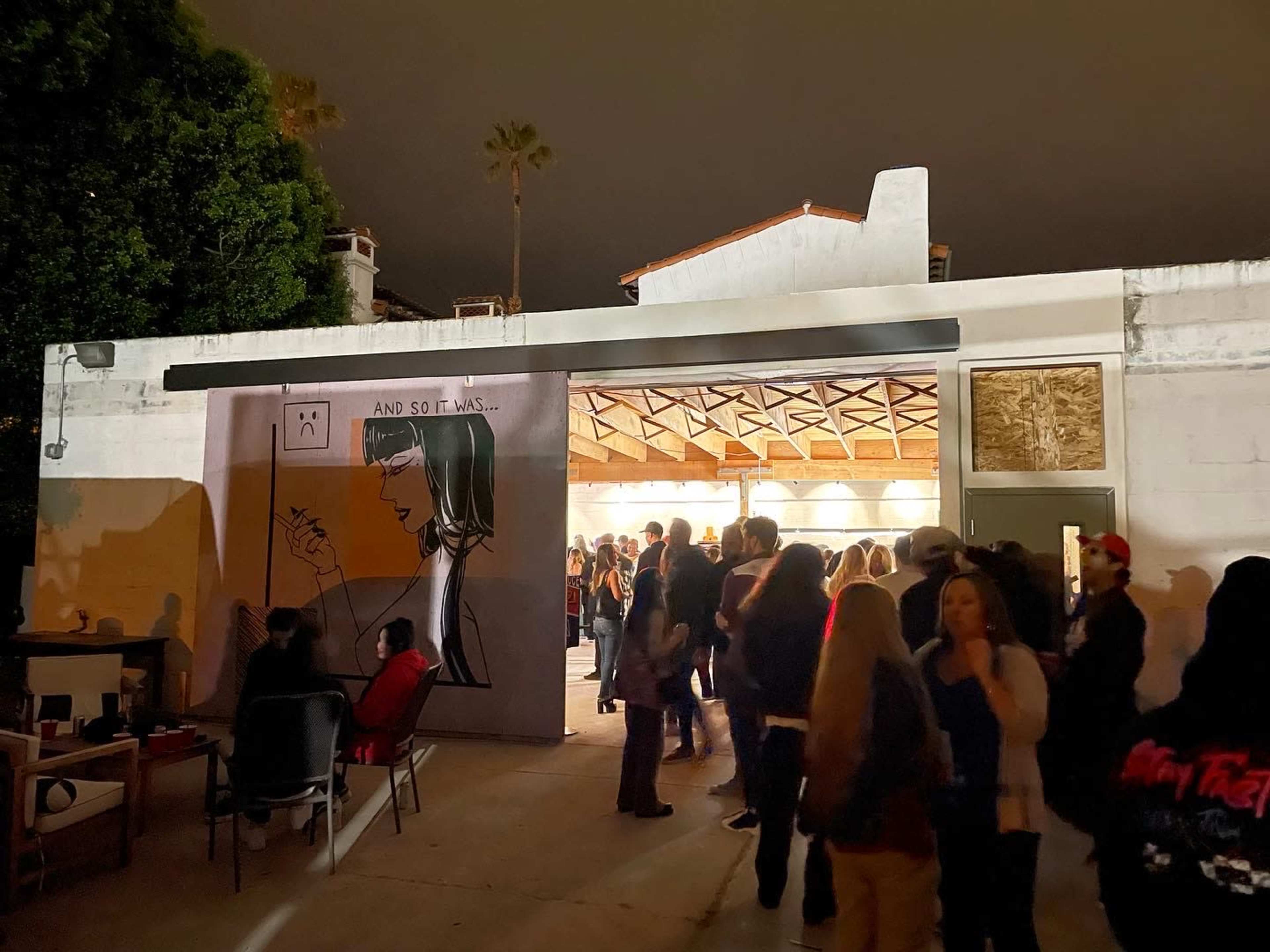 A crowd gathers outside a building featuring a large mural of a woman holding a pencil, under a night sky.