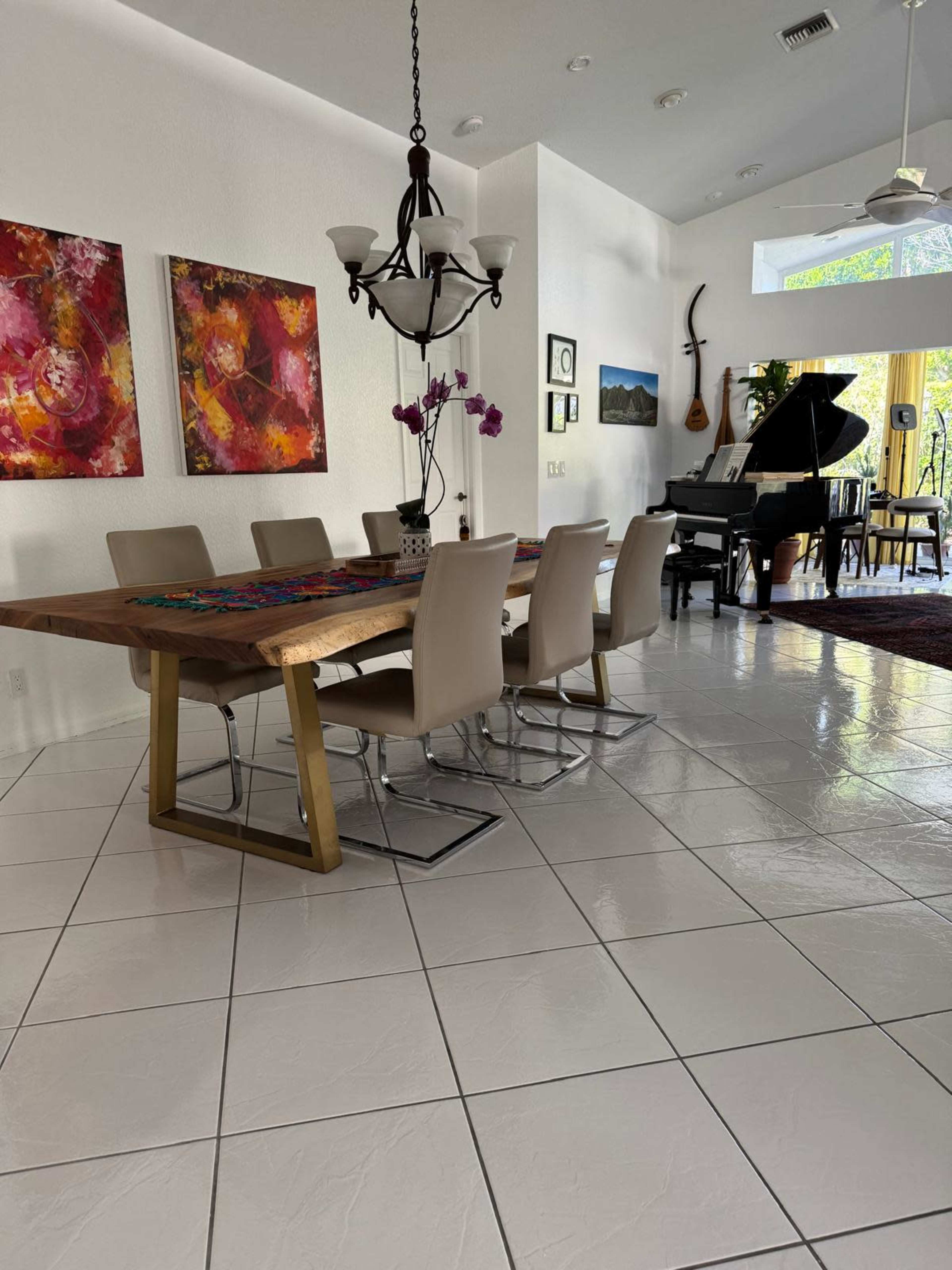 The image shows a dining area with a large wooden table and six beige chairs, adjacent to a piano and adorned with colorful artwork on the walls.