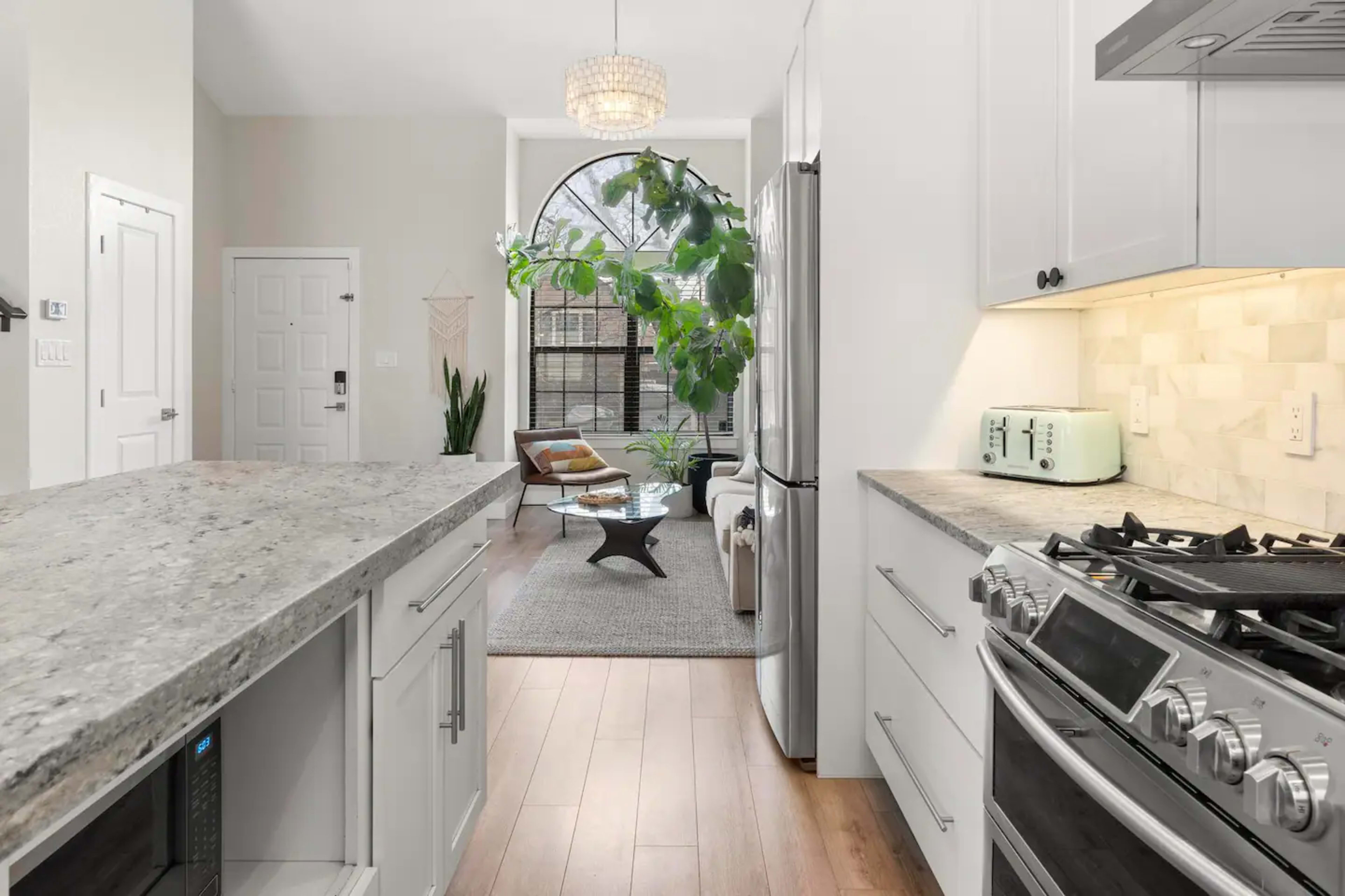 A modern kitchen features light-colored cabinets, a granite countertop, and a stainless steel gas stove, leading into a living area with a large window and indoor plants.