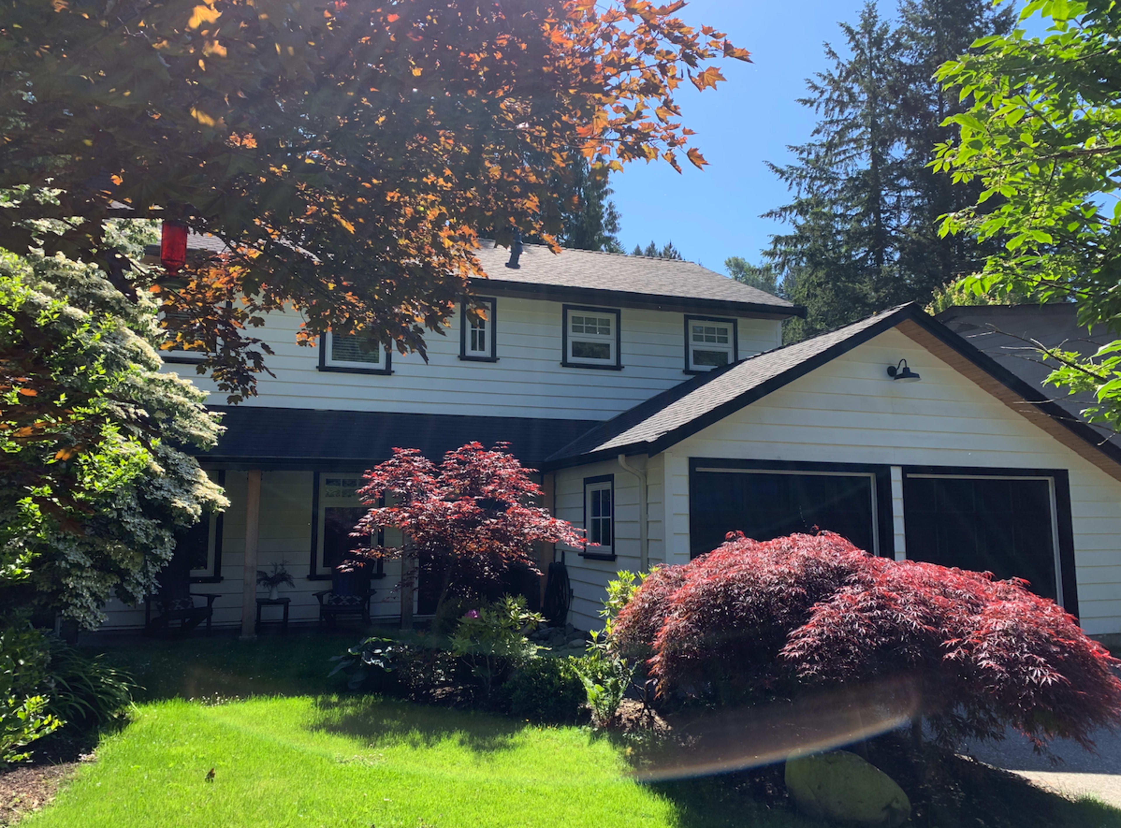 A two-story white house with black trim surrounded by green grass and trees, featuring a red maple tree in the front yard.