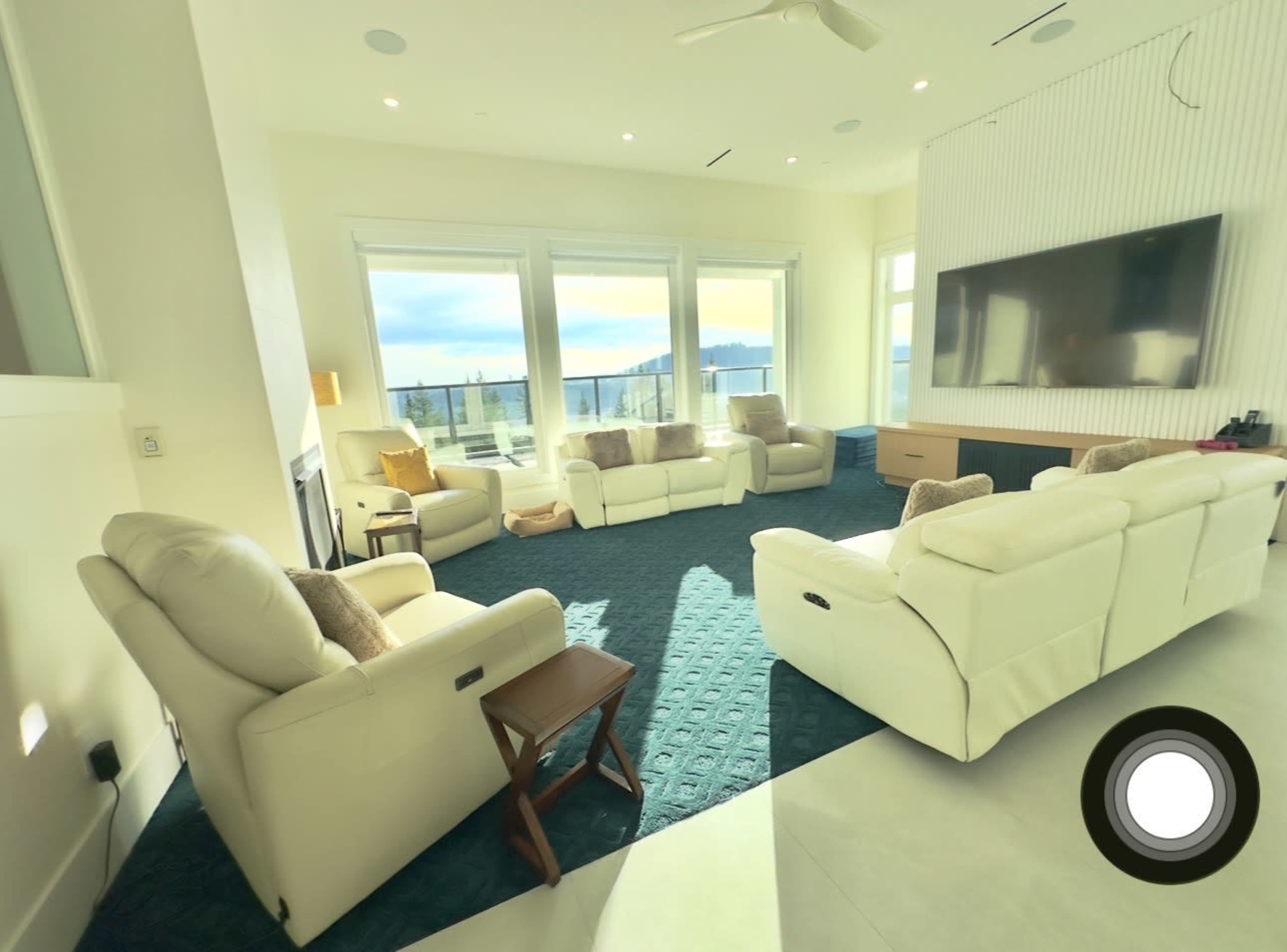 The image shows a modern living room with white leather seating arranged around a blue carpet, large windows, and a wall-mounted television.