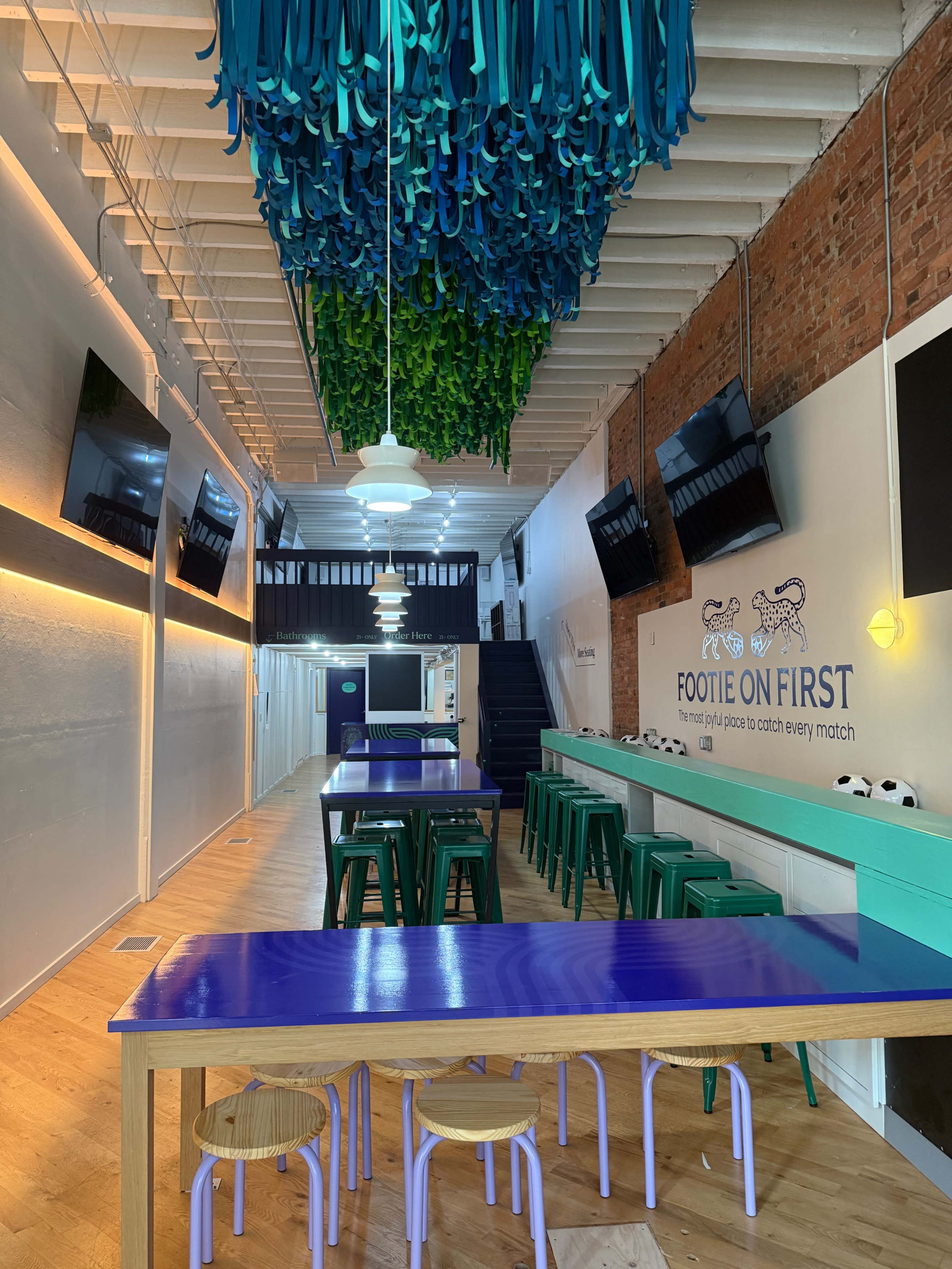 The image shows a spacious, brightly lit room with colorful hanging decorations, modern tables, and multiple screens mounted on the walls.