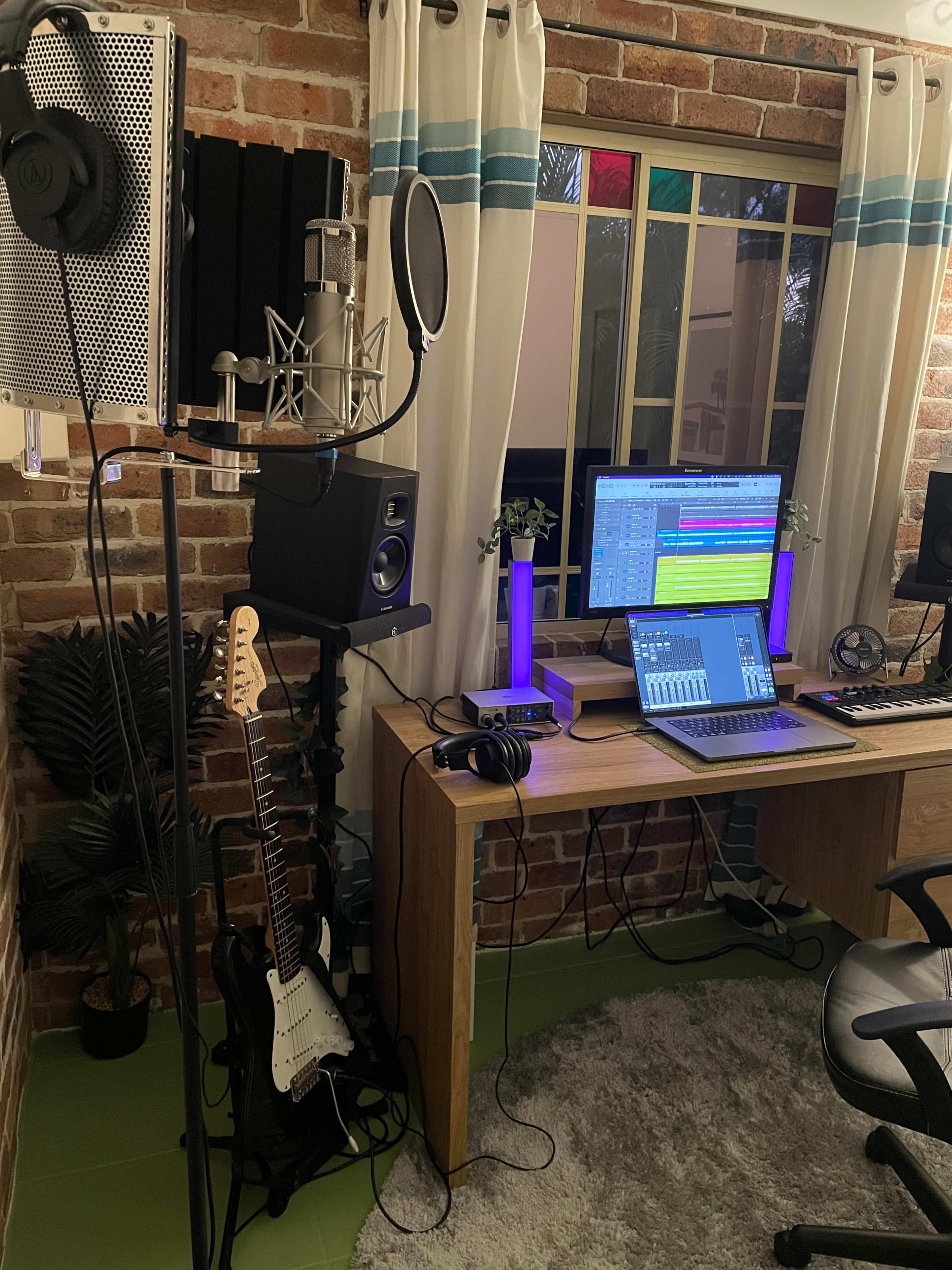 A home music studio features a microphone setup, computer with audio software, and various instruments arranged around a wooden desk.