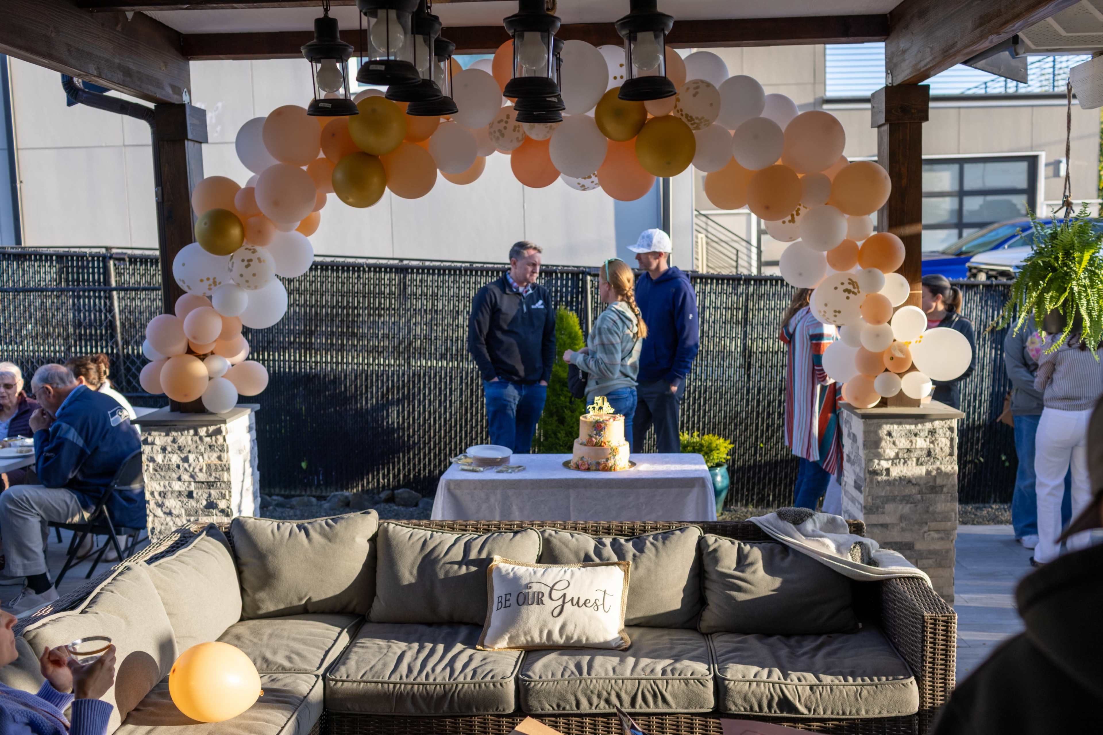A gathering space features a balloon arch and a dessert table, with guests conversing in the background.