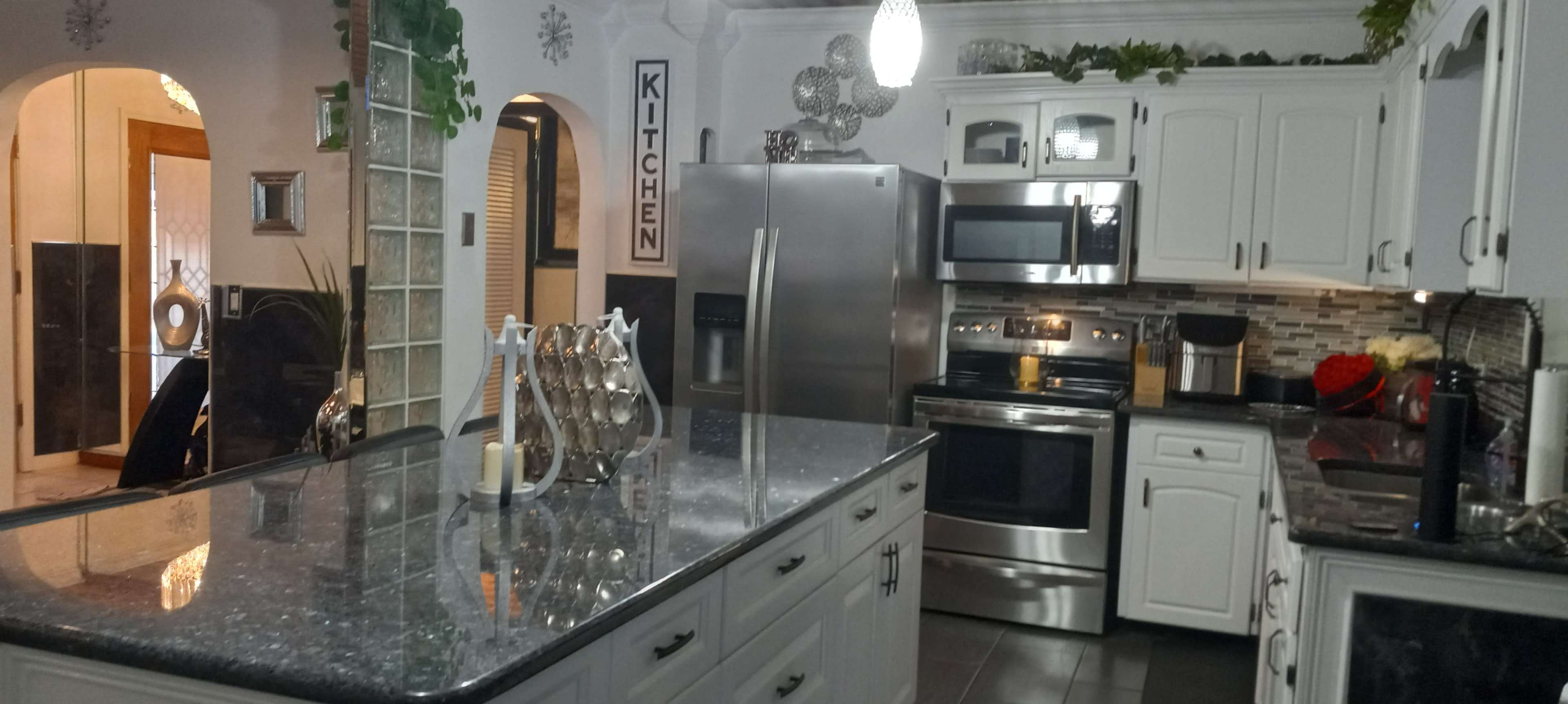 The image shows a modern kitchen with white cabinetry, stainless steel appliances, and a large granite island, illuminated by pendant lighting.